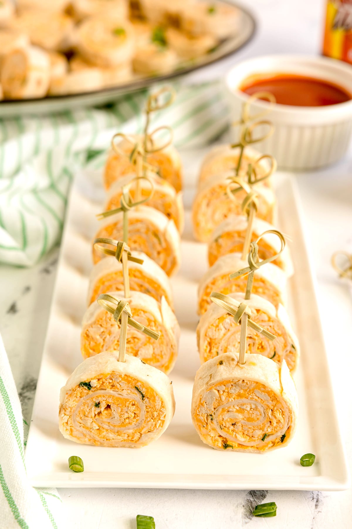 A row of Buffalo Chicken Pinwheels with a creamy orange filling, each secured with a toothpick, arranged on a rectangular white plate. Sauce in a small bowl is visible in the background.