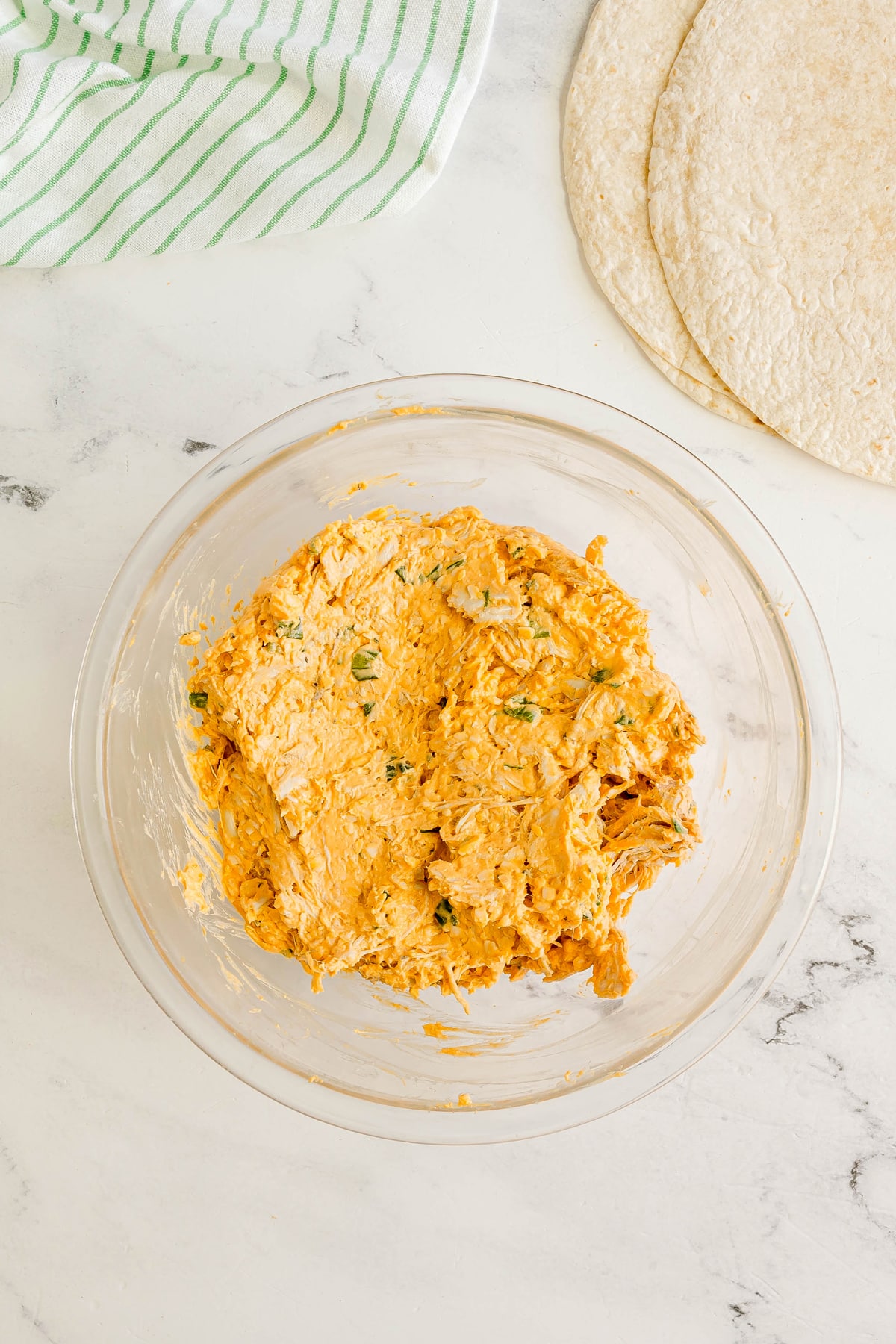 Next step in preparing Buffalo Chicken Pinwheels is to add chicken to creamy, mixture with green flecks. Two flour tortillas and a striped towel are on the white countertop nearby.