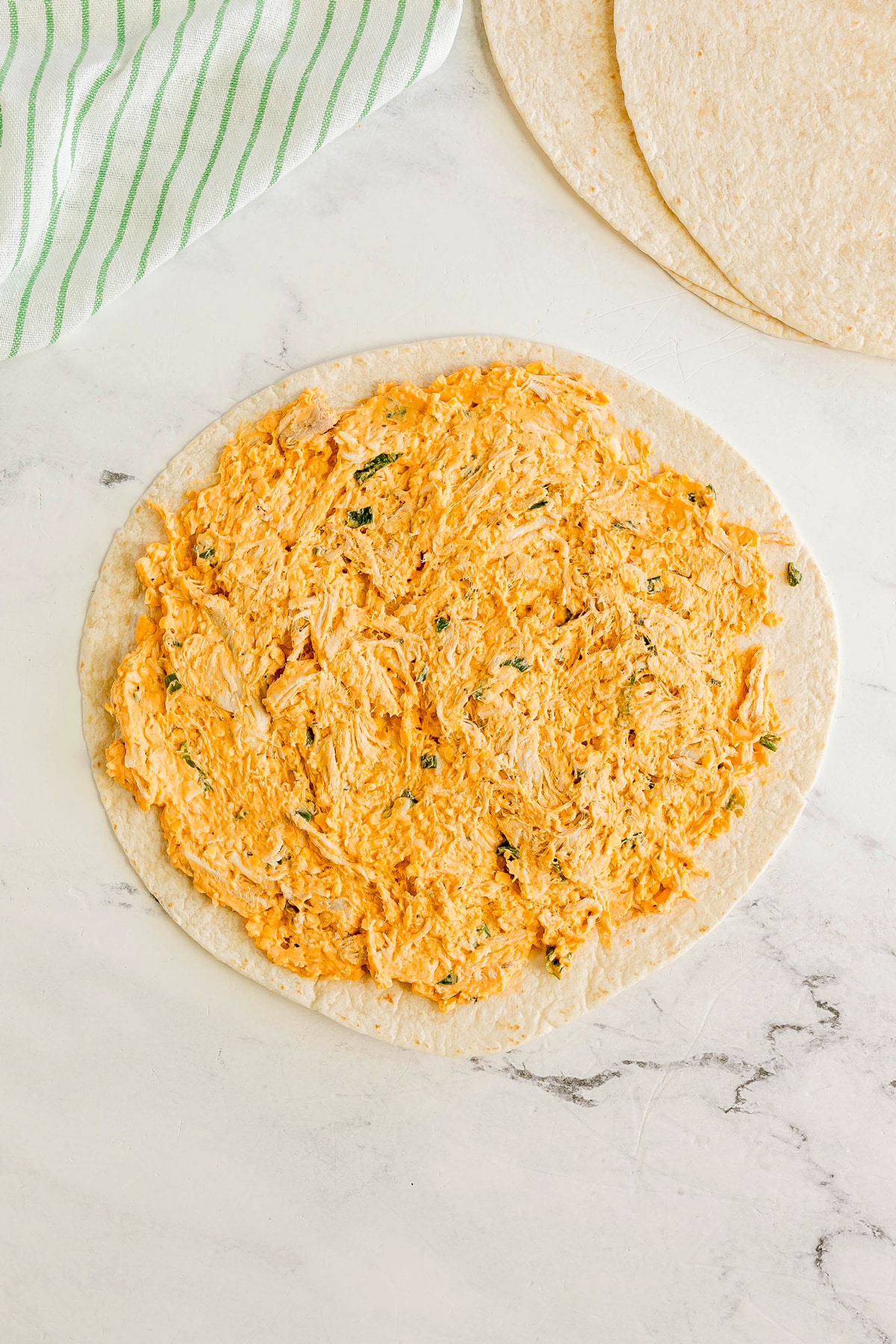 Next step in preparing Buffalo Chicken Pinwheels is to place tortilla on a marble surface is spread with a creamy orange chicken mixture; a green-striped towel and plain tortillas are nearby.