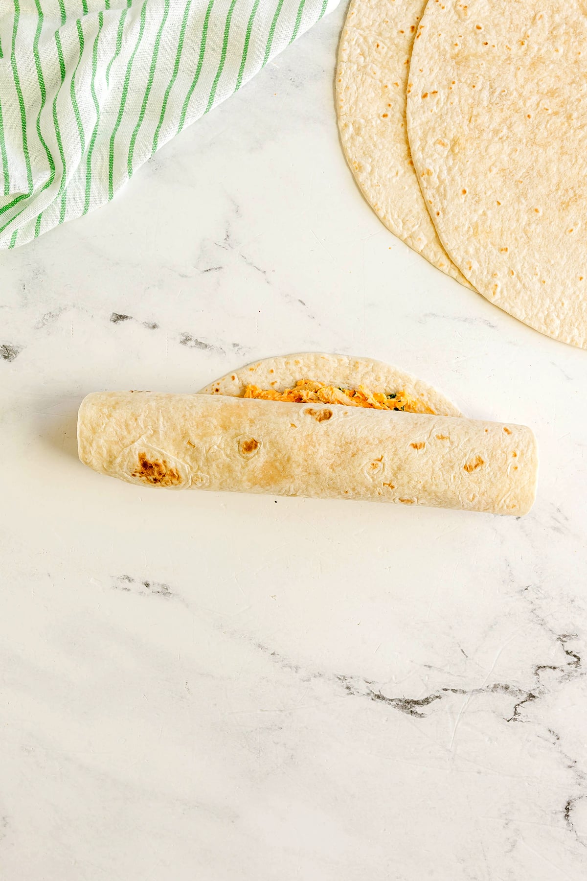 A rolled flour tortilla is placed on a marble countertop, with a flat tortilla and a green-striped kitchen towel nearby. This is one of the step in preparing Buffalo Chicken Pinwheels