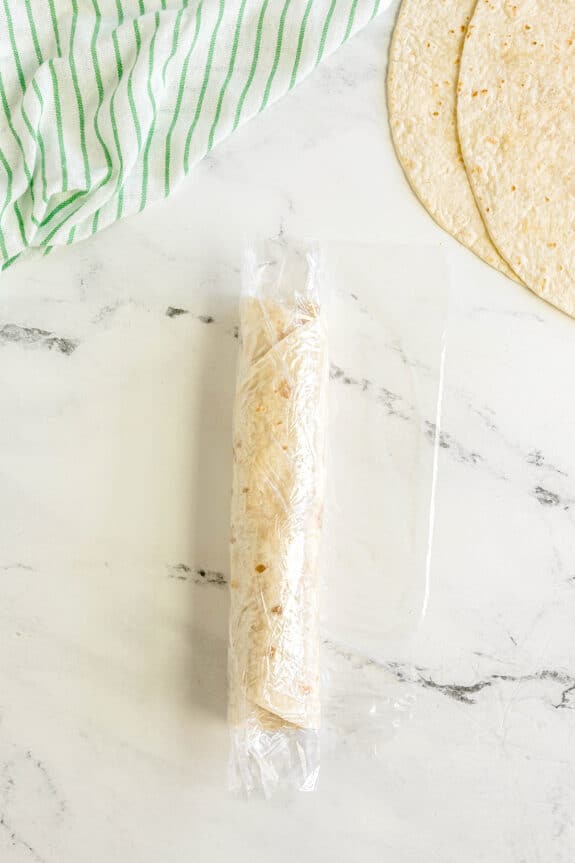 A rolled Buffalo Chicken Pinwheels wrapped in plastic wrap sits on a marble surface next to a green-striped towel and two flat tortillas.