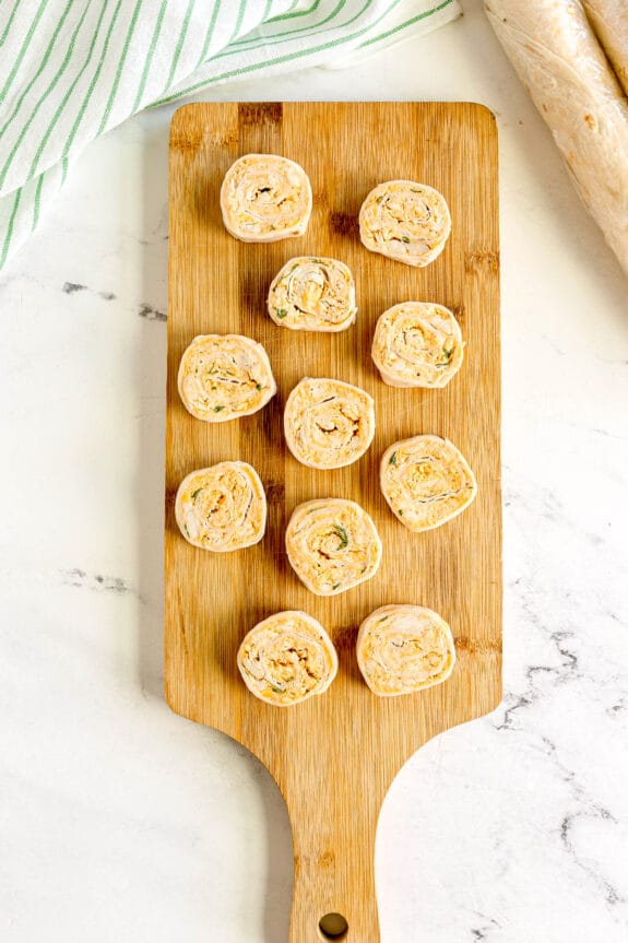 Twelve Buffalo Chicken Pinwheels with a creamy filling are arranged on a wooden cutting board, set on a white surface.