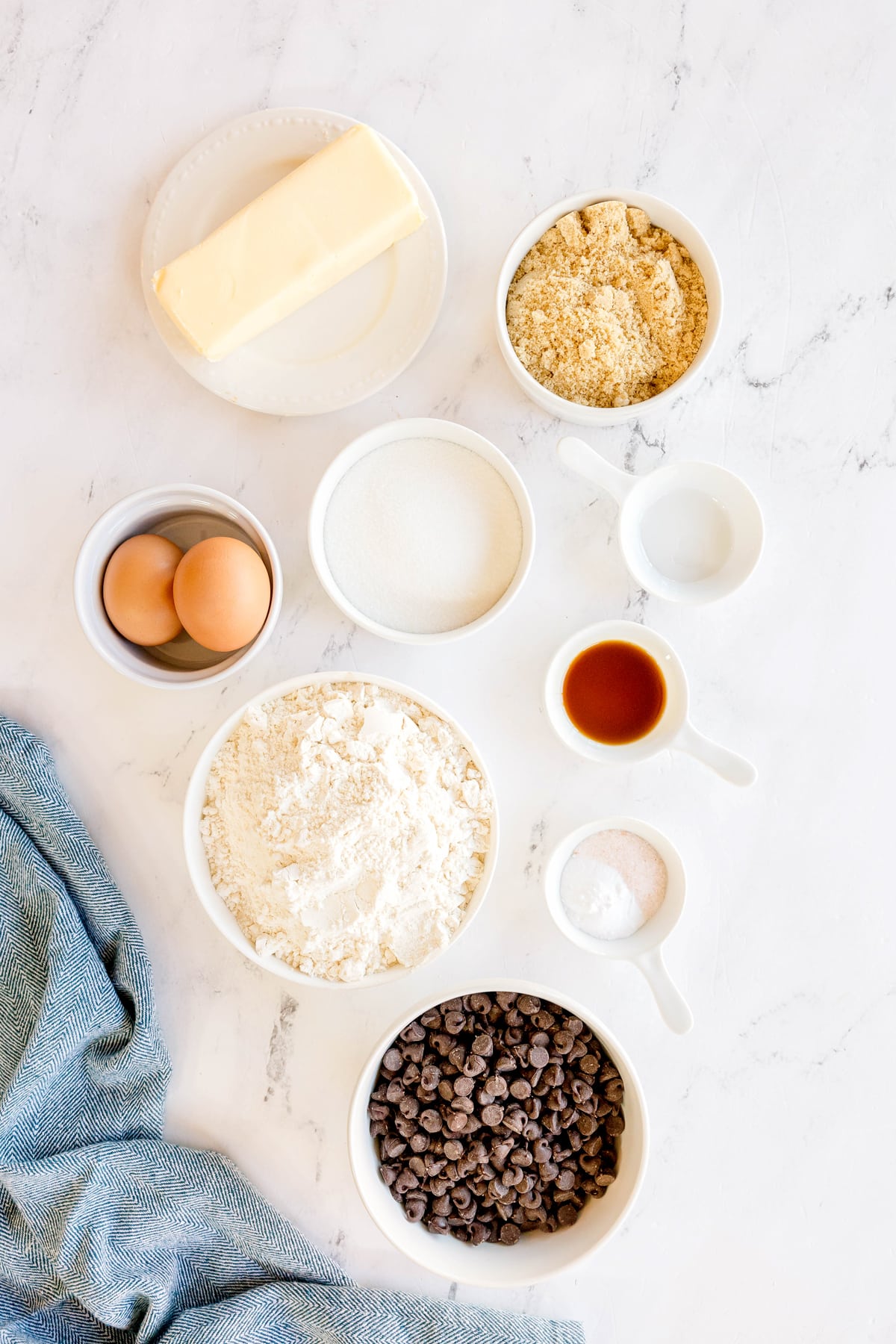 Top-down view of baking ingredients for Chocolate Chip Cookies recipe in bowls on a white surface, including butter- flavored Crisco, brown sugar, granulated white sugar, water, vanilla extract, eggs, all-purpose flour, baking soda, salt, and semi-sweet Chocolate chips