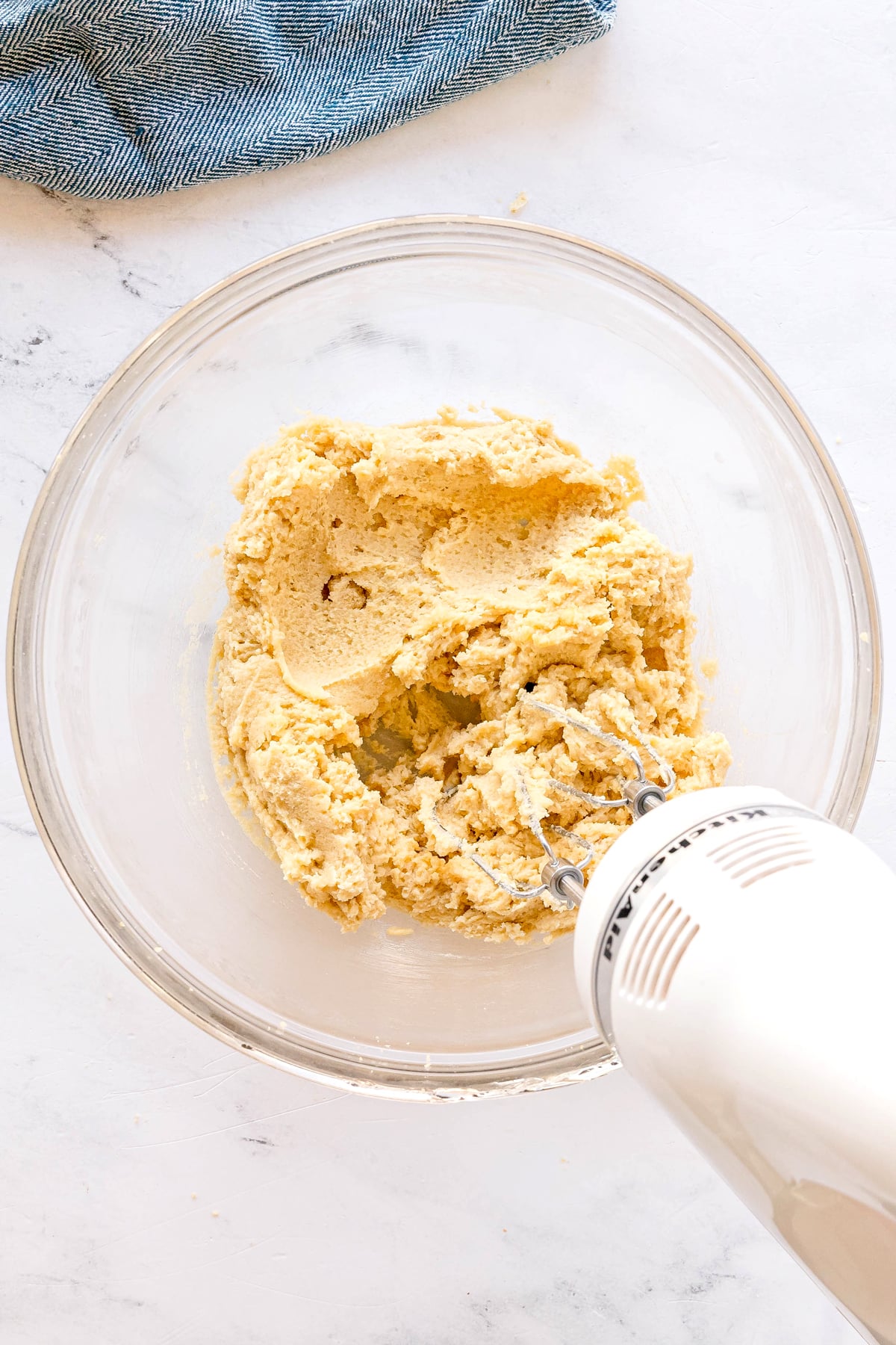 A hand mixer blends cookie dough consists of Crisco, brown sugar, white sugar, water, and vanilla, in a clear glass bowl on a white surface, with a blue cloth visible in the corner for Chocolate Chip Cookies recipe