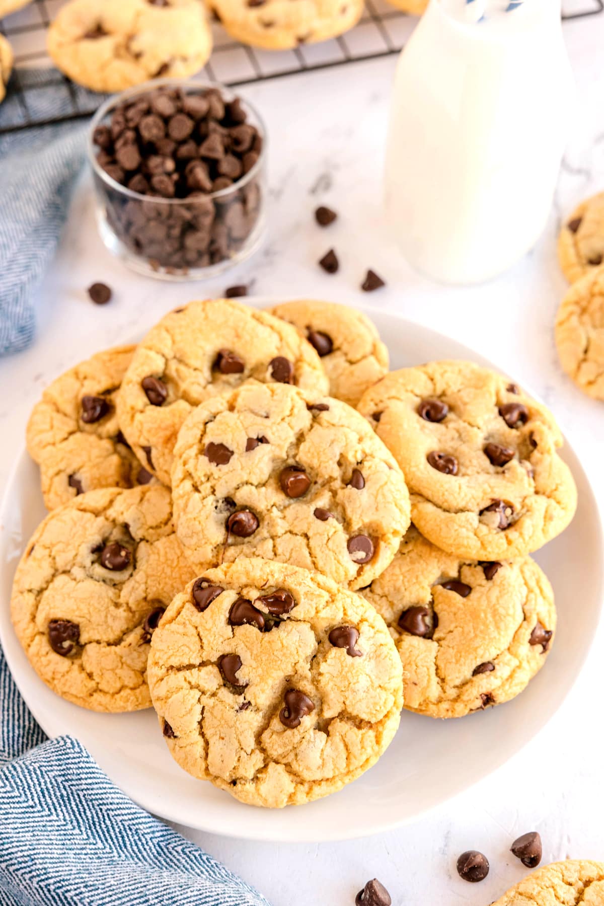 A plate of chocolate chip cookies sits on a white surface, with a glass of chocolate chips, a milk bottle, and a cooling rack with more cookies in the background.