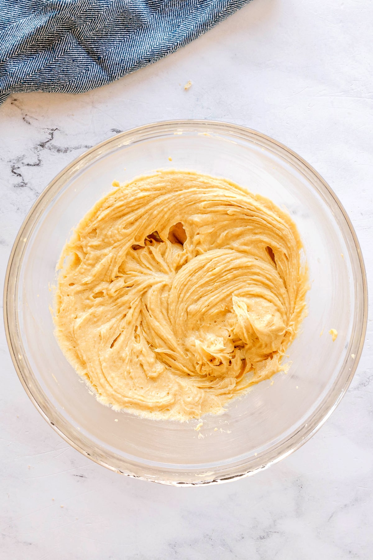 A glass bowl filled with creamy, swirled yellow batter for Chocolate Chip Cookies on a light surface next to a blue cloth.