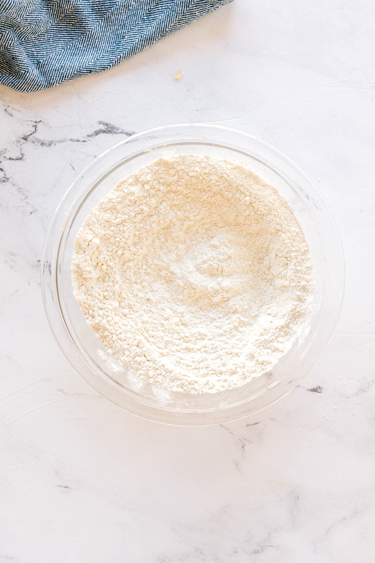 A clear glass bowl filled with flour, baking soda, and salt mixture, sits on a white marble surface, with part of a blue fabric visible in the corner for Chocolate Chip Cookies recipe/