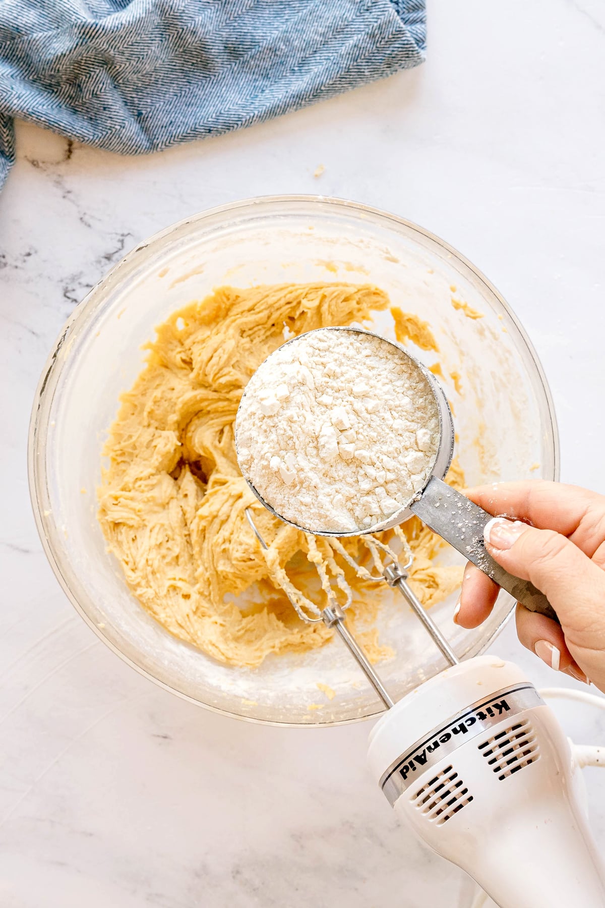A person adds a cup of flour mixture to a bowl of Chocolate Chip Cookie dough mixture while using a hand mixer on a marble countertop.