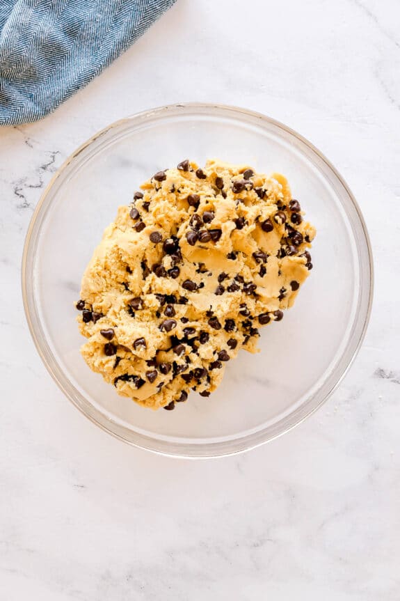 A glass bowl filled with raw Chocolate Chip Cookie dough sits on a white marble surface.