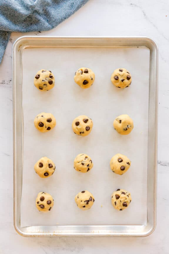 A baking sheet lined with parchment paper holds twelve evenly spaced balls of Chocolate Chip Cookie dough, ready to be baked. A blue cloth is partially visible in the top corner.