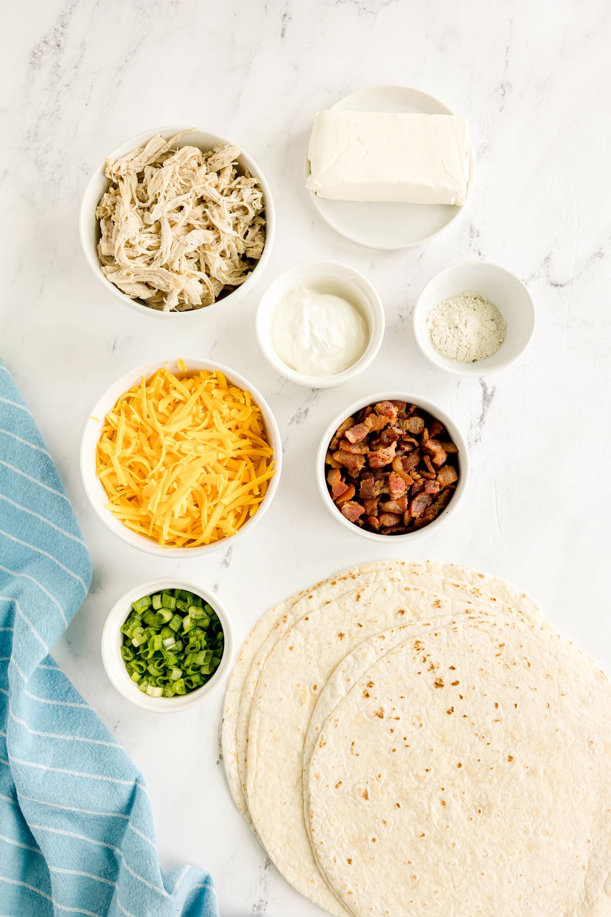 Flat lay of Easy Crack Chicken Pinwheels ingredients are the following: shredded chicken, cream cheese, shredded cheddar, sour cream, ranch seasoning, cooked bacon pieces, chopped green onions, and flour tortillas on a marble surface with a blue towel.
