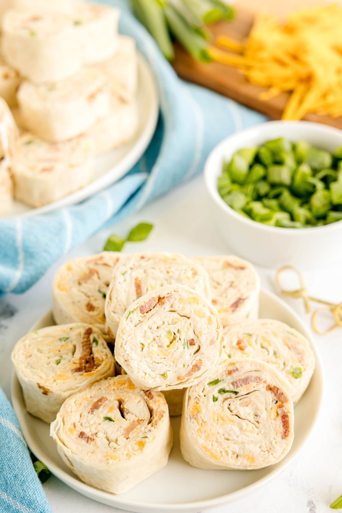 A plate of tortilla pinwheels filled with a creamy mixture, surrounded by a bowl of chopped green onions and shredded cheese in the background.