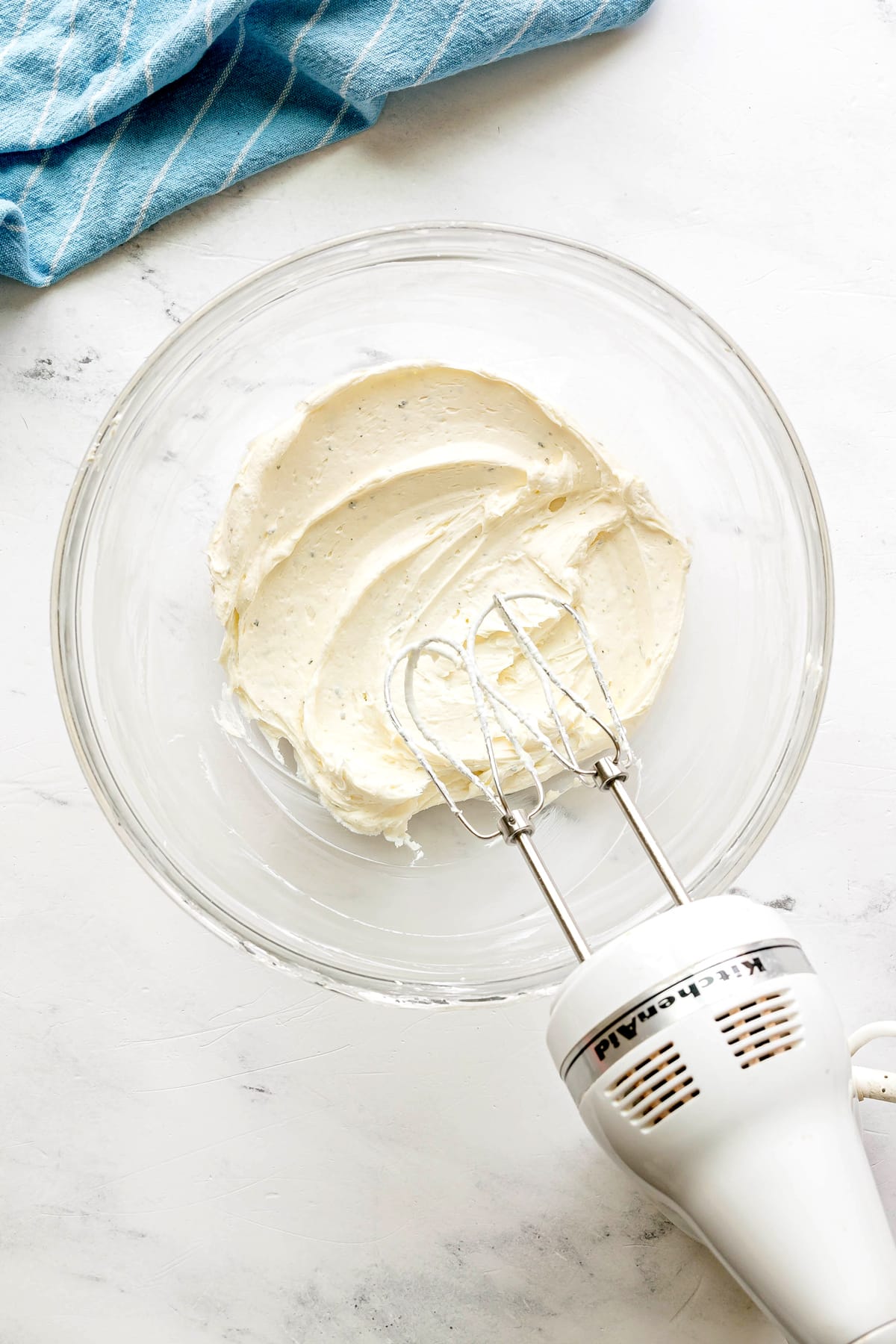 A glass bowl with whipped cream cheese mixture and an electric hand mixer resting inside, on a white surface with a blue striped cloth nearby. This is one of the steps in preparing Easy Crack Chicken Pinwheels