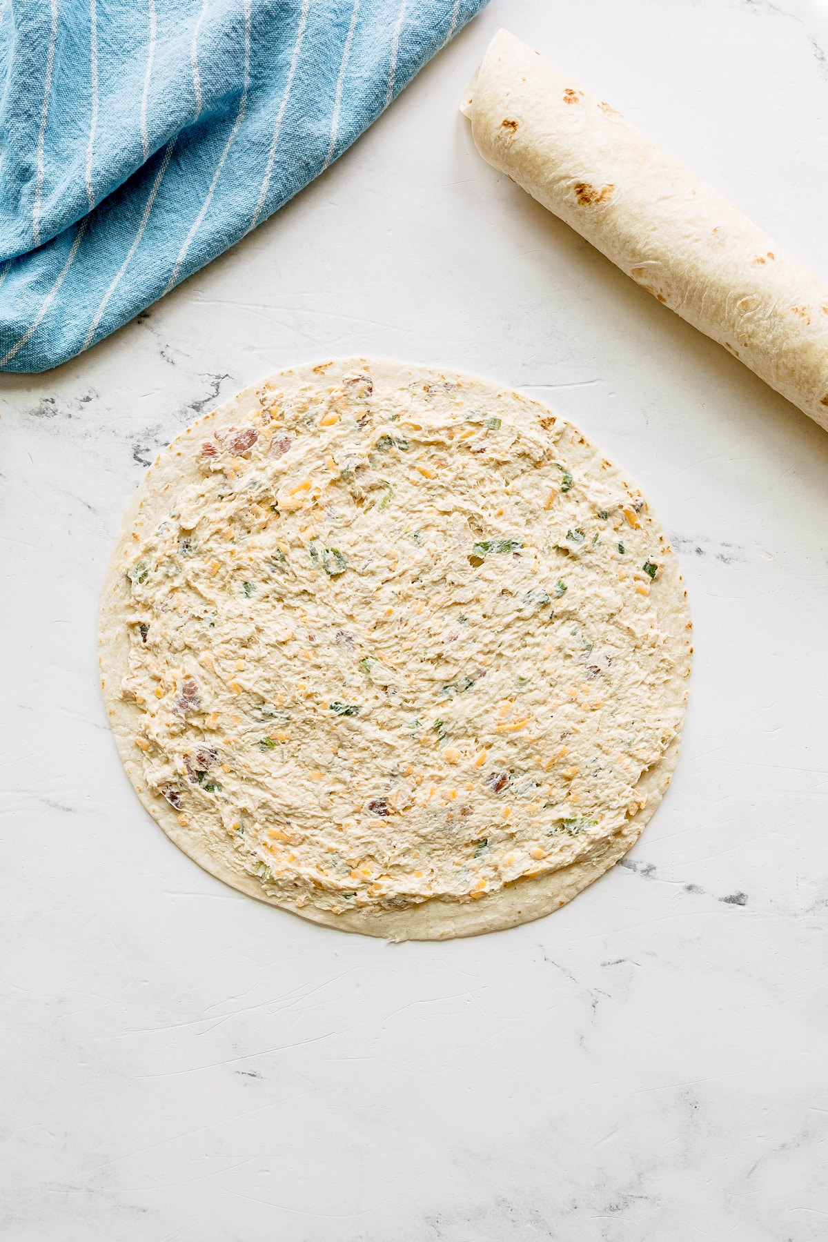 Next step in preparing Easy Crack Chicken Pinwheels is to use tortilla and add spread with a creamy mixture containing herbs and small bits of vegetables, next to a rolled-up tortilla and a blue striped cloth.