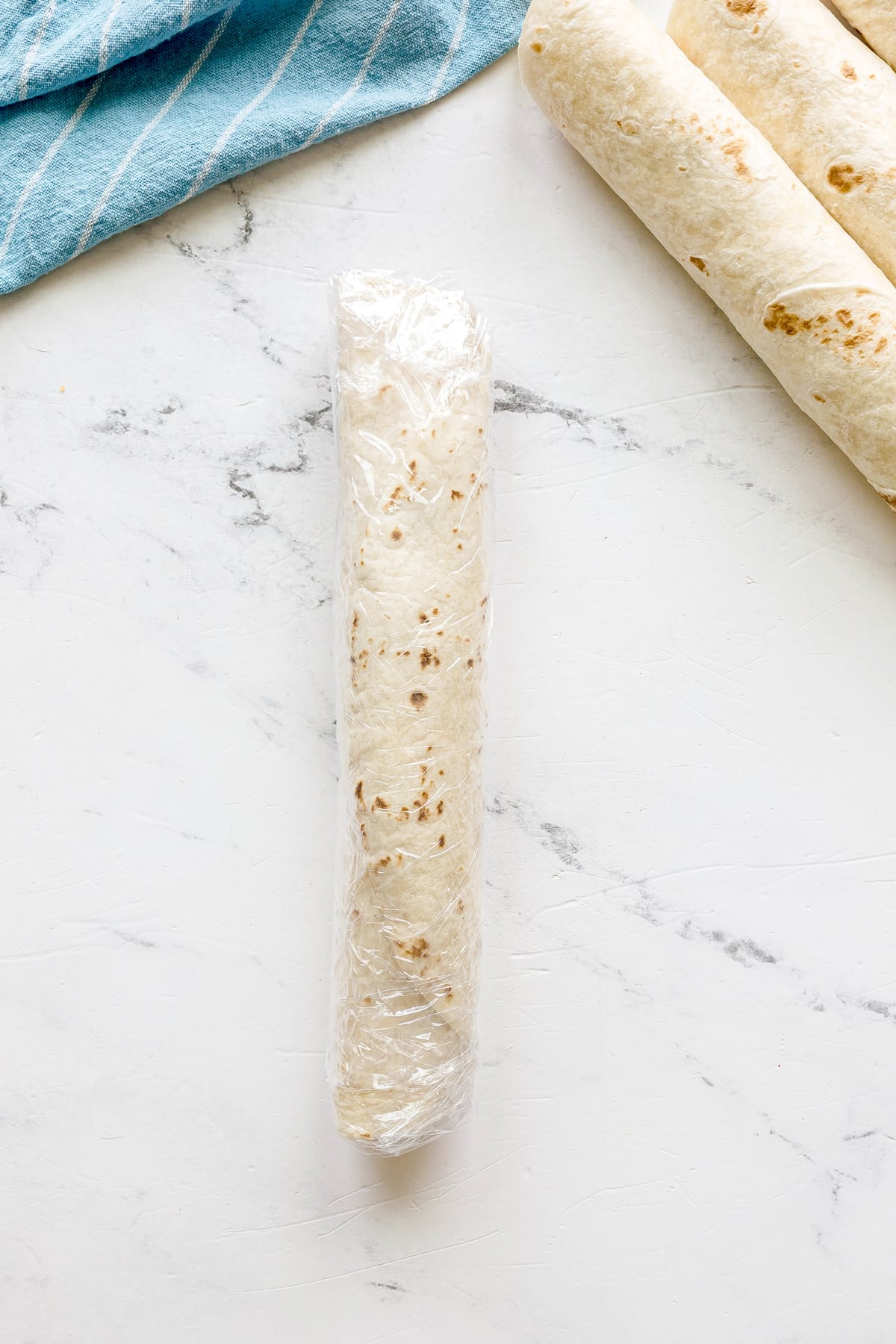 Easy Crack Chicken Pinwheels wrap tightly rolled and covered in plastic wrap is placed on a white marble surface near two unwrapped tortillas and a blue-striped cloth.