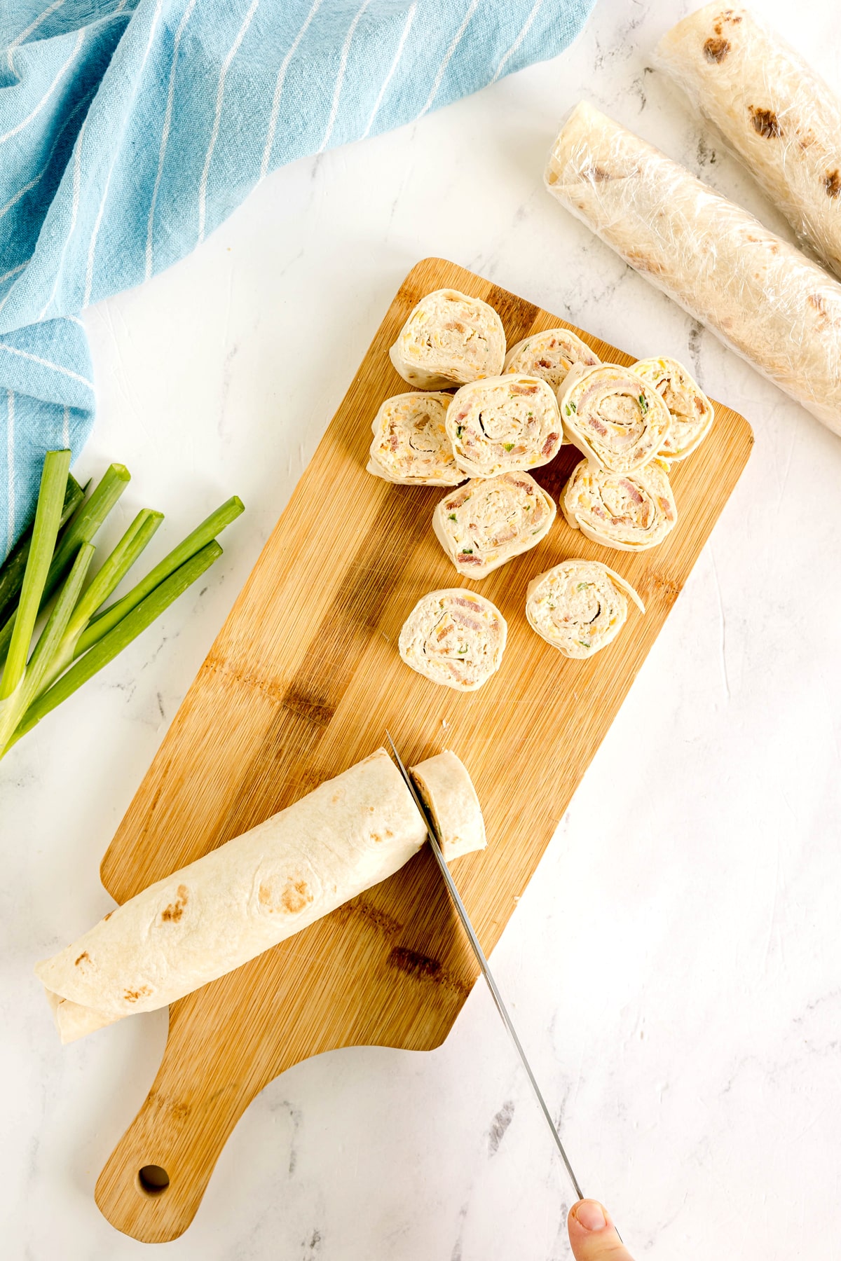 A hand slices a Easy Crack Chicken Pinwheels into bite-sizes on a wooden cutting board, with green onions and more roll-ups nearby.
