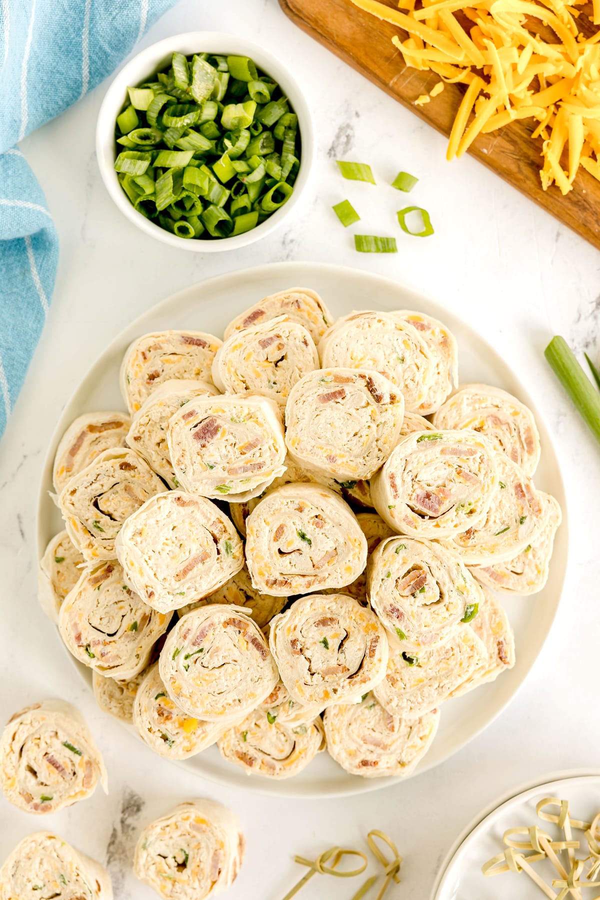 A plate of Easy Crack Chicken Pinwheels filled with a creamy mixture, surrounded by chopped green onions, shredded cheese, and a blue towel on a white surface.