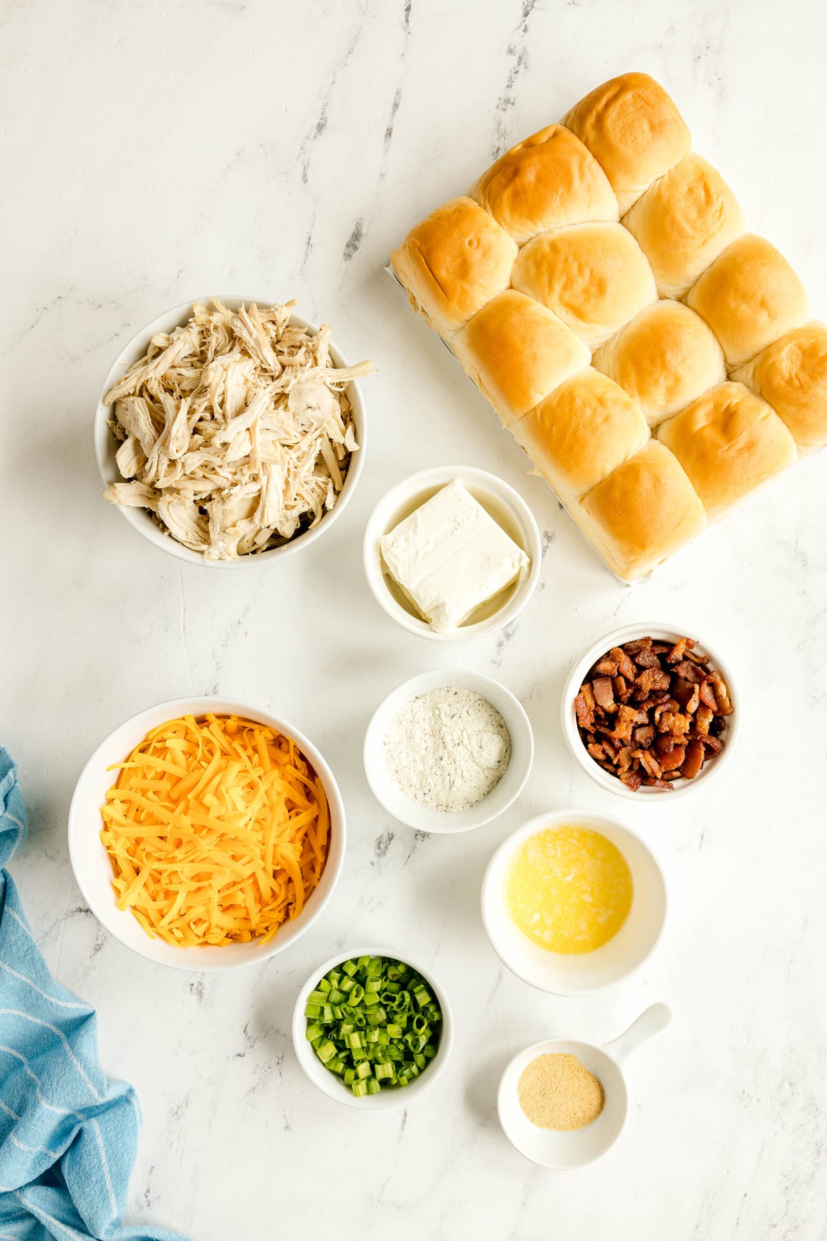 Bowls of Crack Chicken Sliders ingredients include Hawaiian sweet rolls, shredded chicken, sharp cheddar cheese, cream cheese, garlic powder, cooked crumbled bacon, unsalted butter and green onions are arranged on a marbled surface.