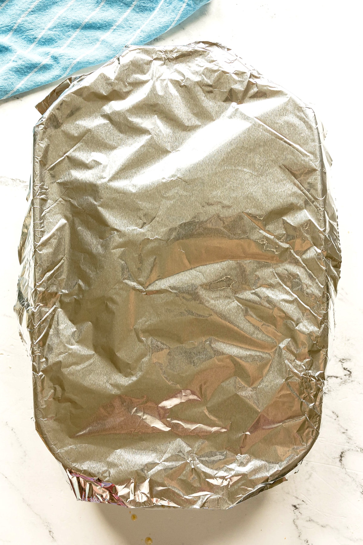 A rectangular baking dish covered with crinkled aluminum foil sits on a white surface near a blue cloth. This is one of the processes in preparing Crack Chicken Sliders