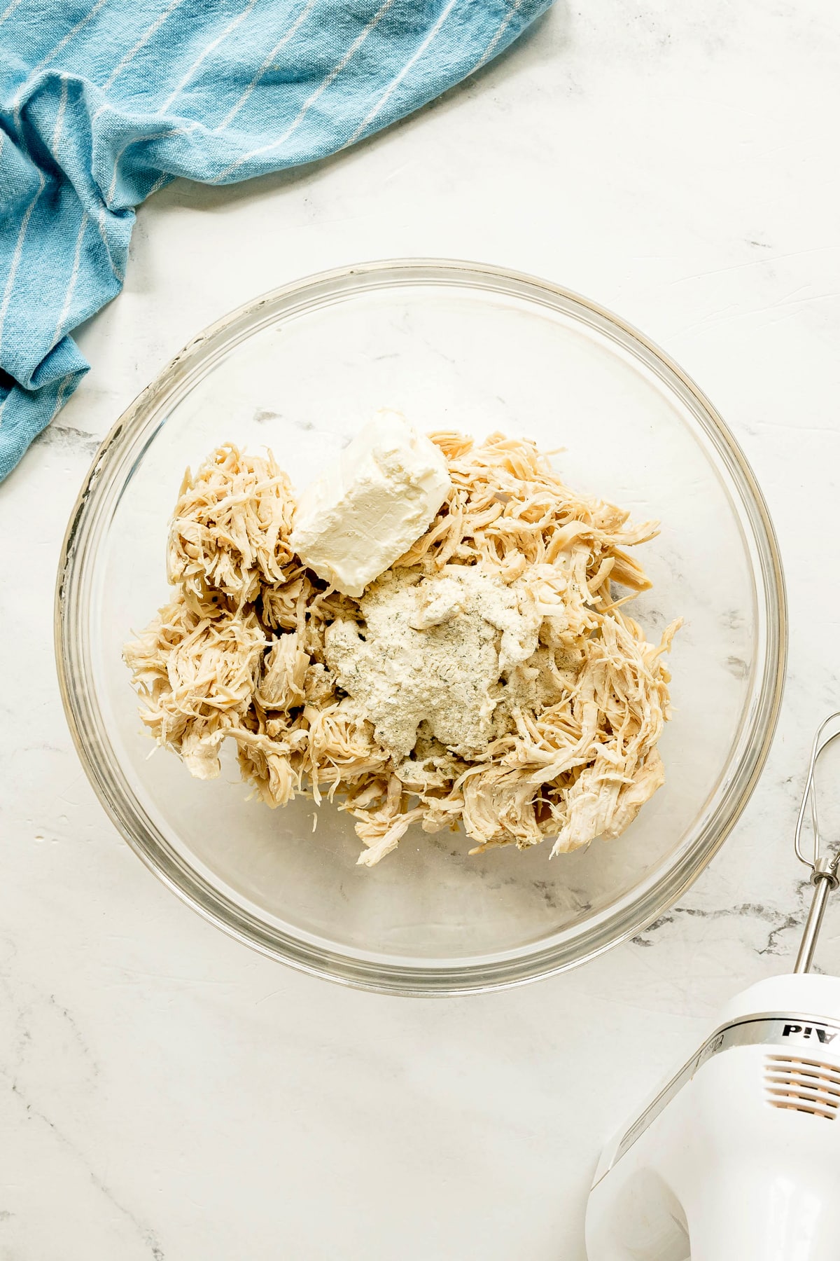 Next step in preparing Crack Chicken Sliders is to use a glass bowl containing shredded chicken, a block of cream cheese, and a powdered seasoning mix on a white countertop with a hand mixer and blue towel nearby.
