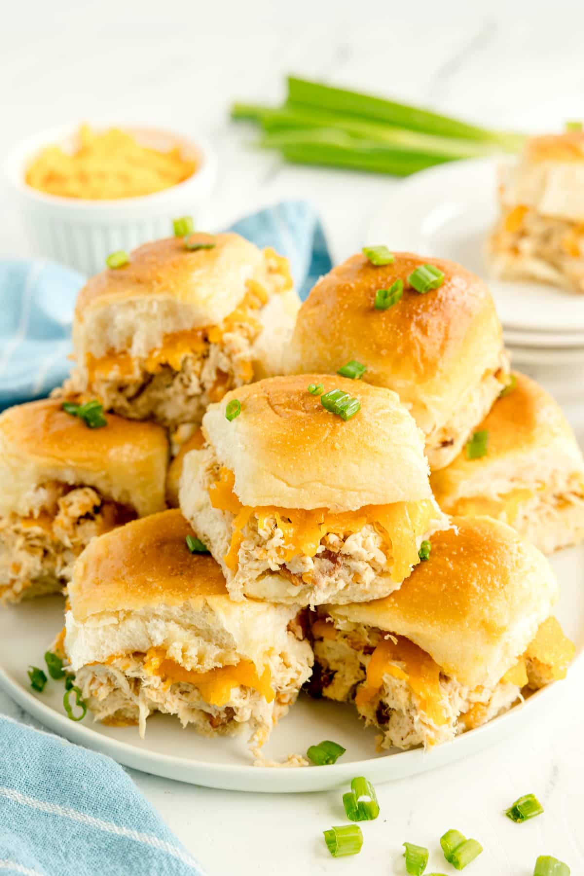 A plate of Crack Chicken Sliders on soft buns, topped with chopped green onions, with a small bowl of dip and a stack of sliders in the background.