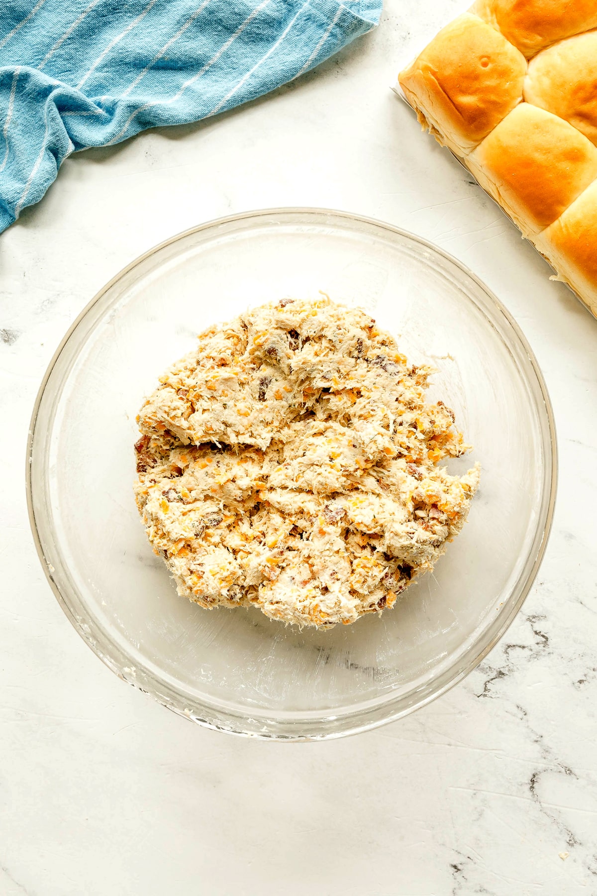 A glass bowl containing mixture with added crumbled bacon and 1 cup of the shredded cheddar cheese is the next step in preparing Crack Chicken Sliders. This sits on a white countertop next to a blue cloth and a tray of baked bread rolls.