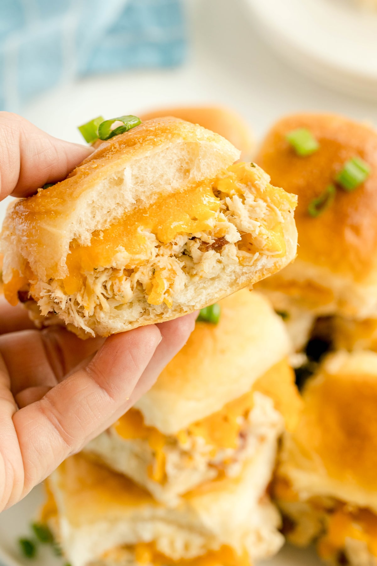 A hand holds a Crack Chicken Slider filled with shredded chicken and melted cheddar cheese; more sliders are stacked on a plate below.