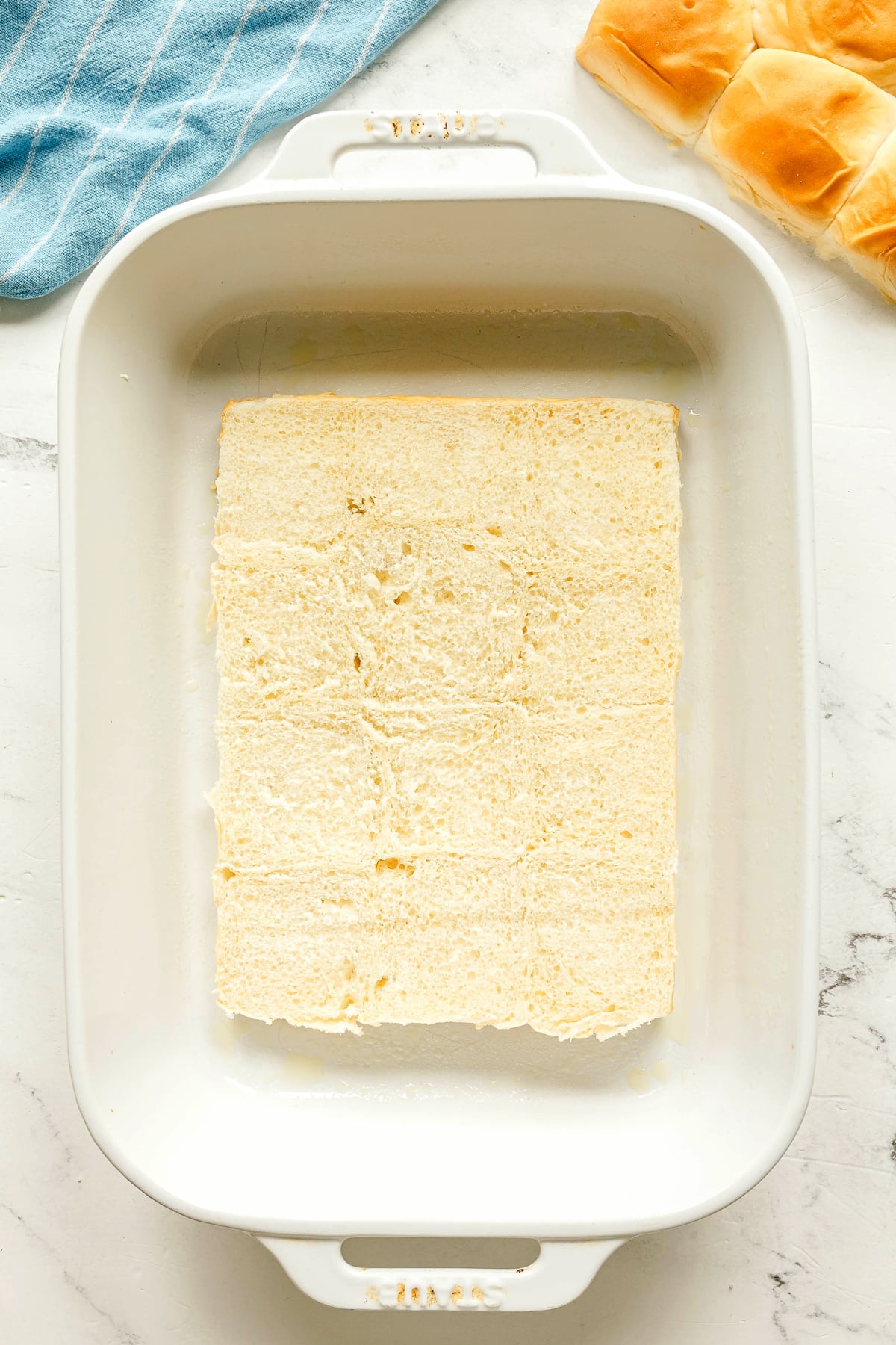 White baking dish with a single layer of sliced bread rolls placed inside; extra rolls and a blue cloth are visible at the top edge. This is for Crack Chicken Sliders recipe