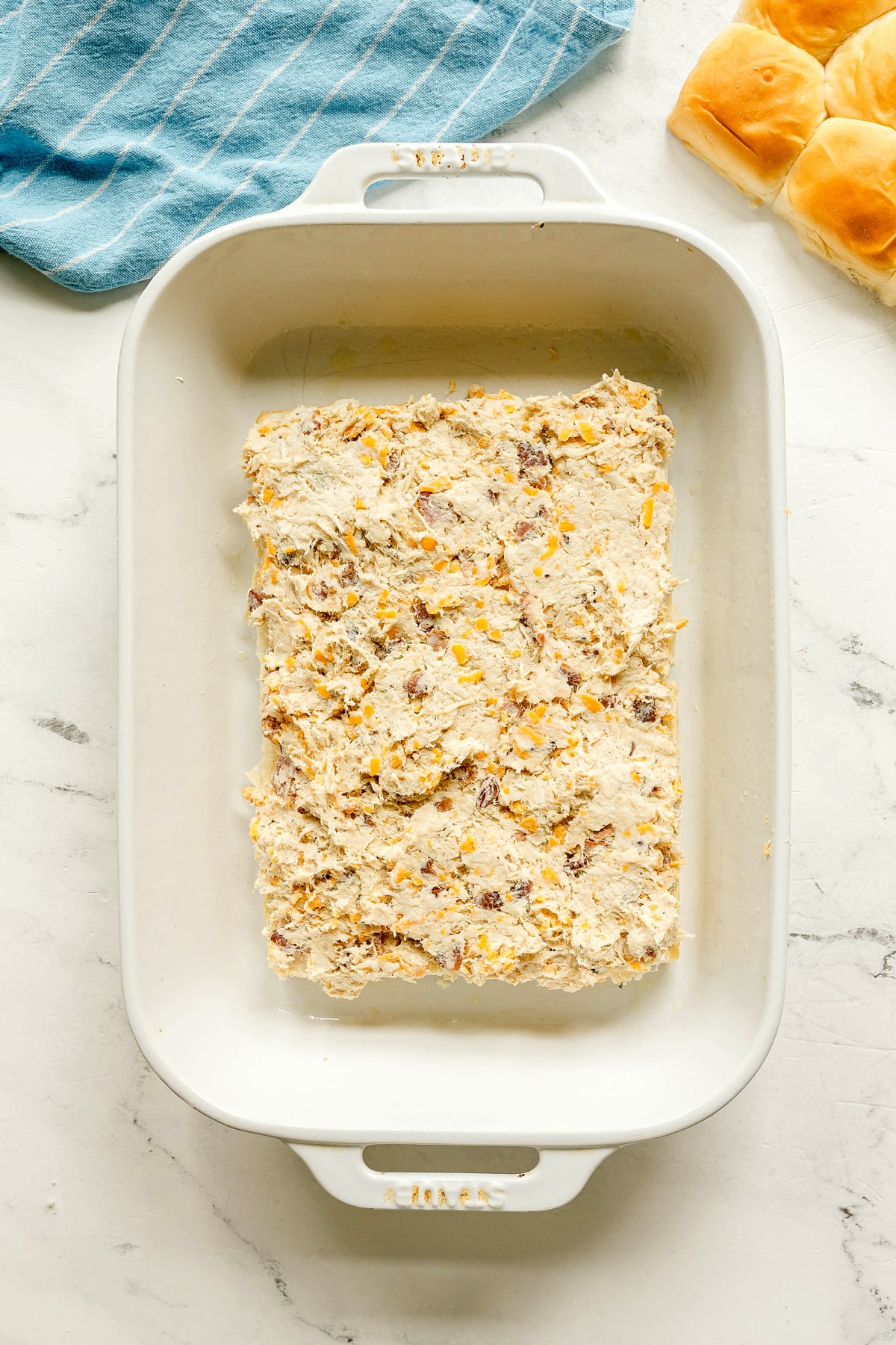 Next step in preparing Crack Chicken Sliders is to spread unbaked mixture containing shredded cheese and pieces of meat, on a marble surface next to a blue towel and dinner rolls.
