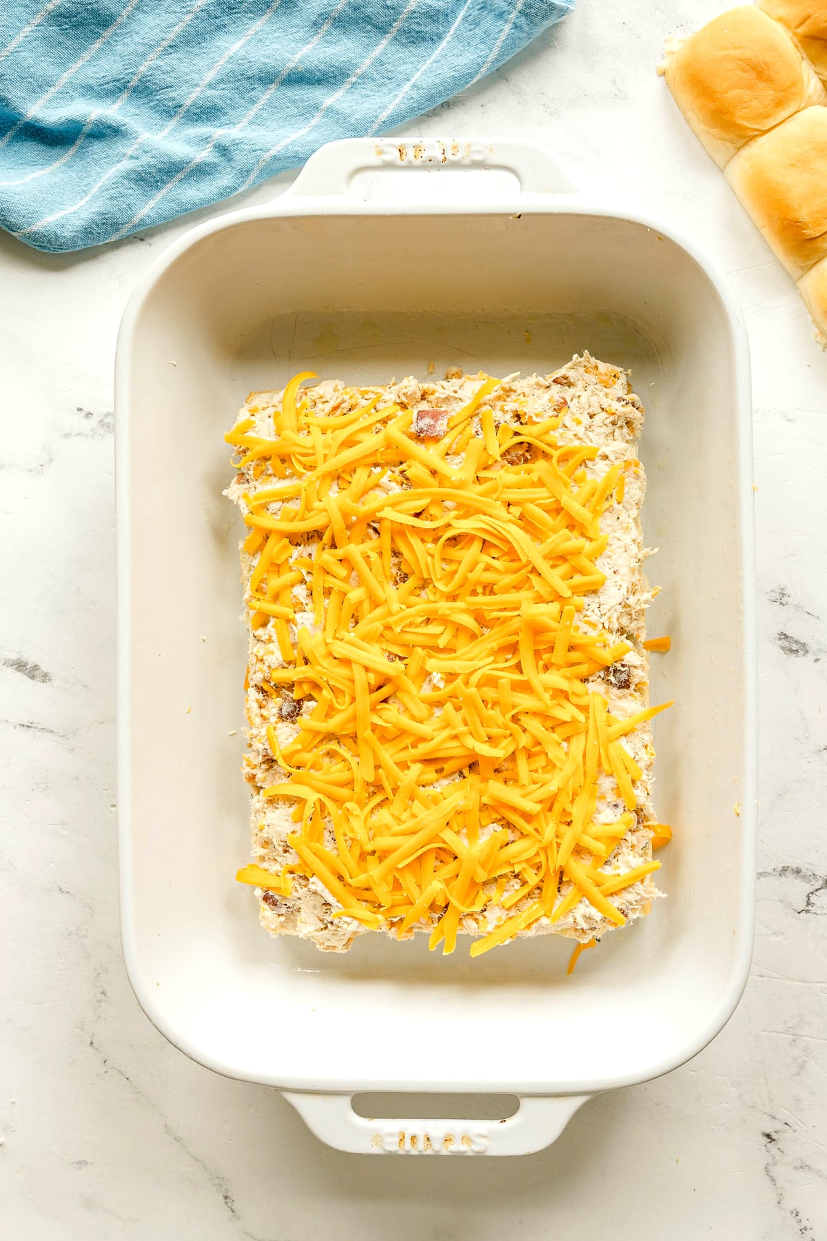 A white baking dish for Crack Chicken Sliders recipe is filled with a creamy mixture topped with shredded cheddar cheese, placed on a marble surface next to a blue towel and bread rolls.