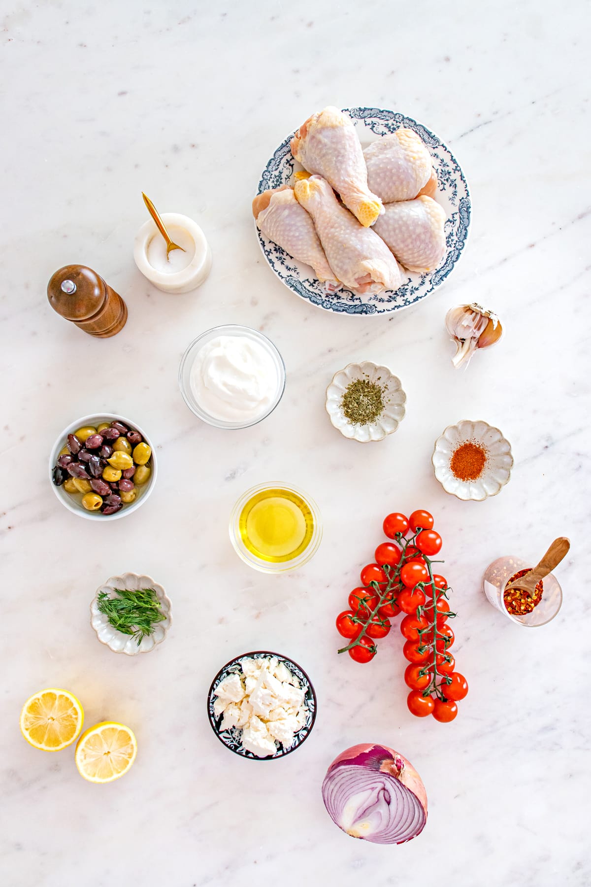 Assorted ingredients for One Pan Greek Chicken Drumsticks on a marble surface, including raw chicken drumsticks, plain Greek yogurt, Olive oil, lemon juice, garlic, red pepper flakes, salt, black pepper, Italian seasoning, paprika, and fresh dill.
