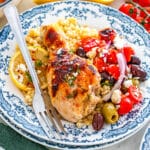 A plate of One Pan Greek Chicken Drumsticks with roasted chicken, couscous, lemon slice, and a side of tomato, olive, onion, and feta salad; a fork is placed on the plate.