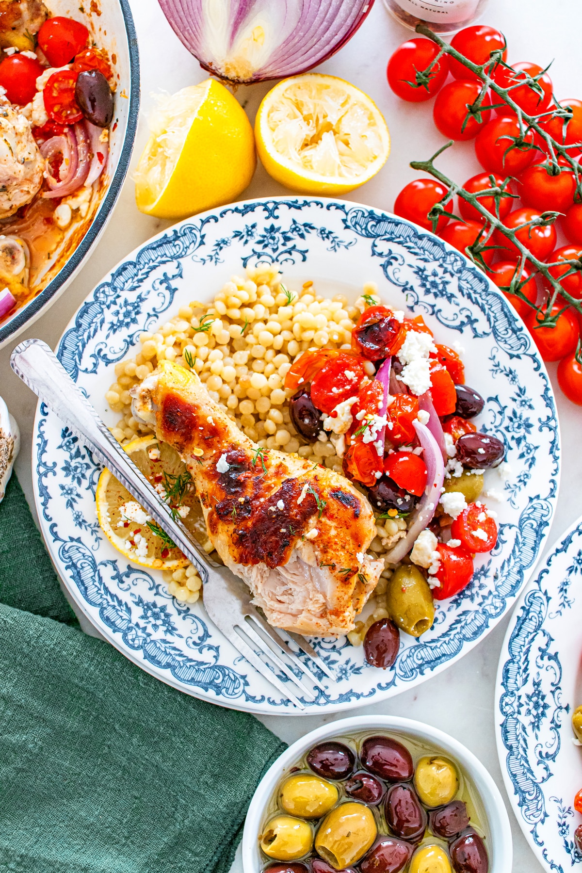 A plate with One Pan Greek Chicken Drumstick, couscous, and a salad of tomatoes, red onion, feta, and olives, garnished with a lemon wedge. Cherry tomatoes and mixed olives are nearby.