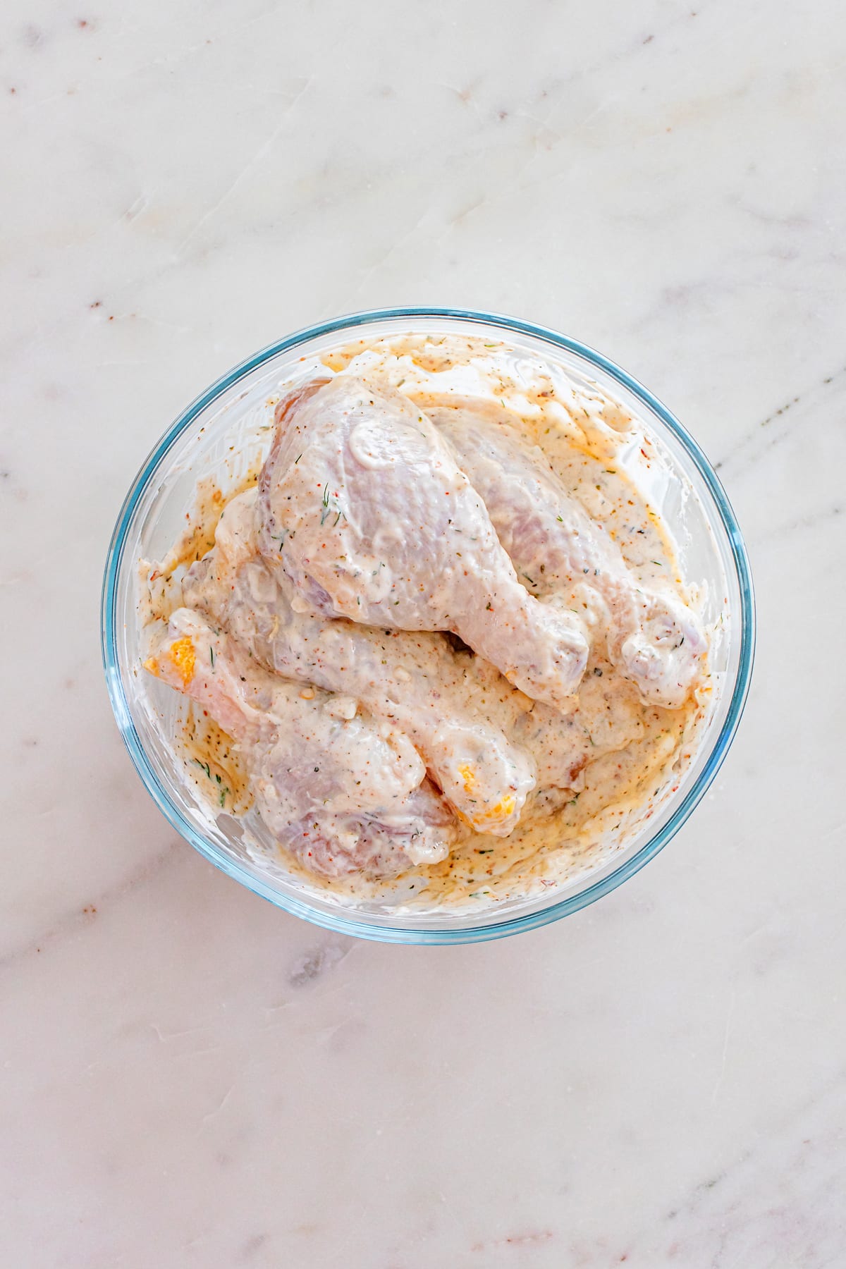 Next step in preparing One Pan Greek Chicken Drumsticks is to coat raw chicken drumsticks in a creamy marinade with herbs, placed in a clear glass bowl on a light marble surface.
