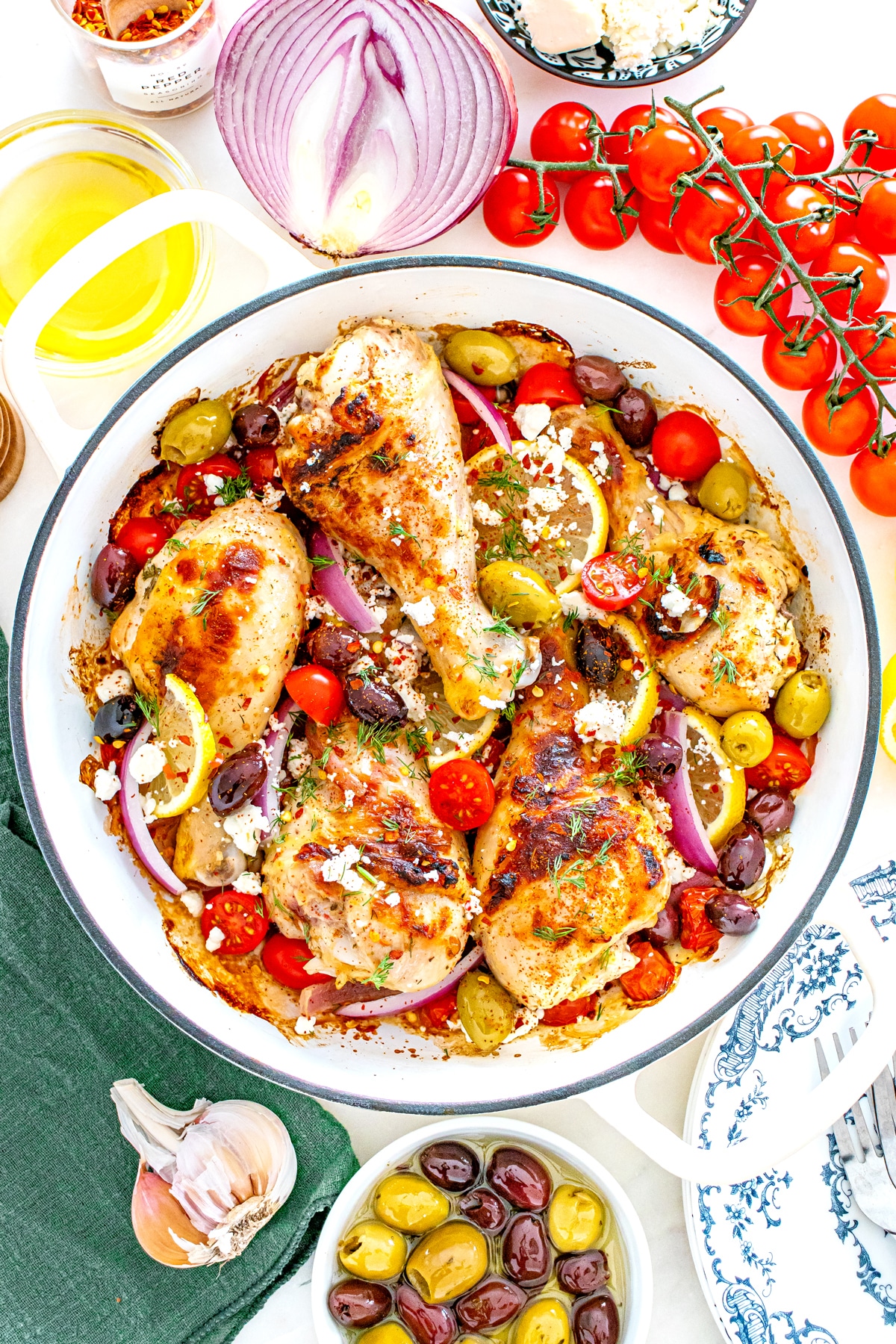 Freshly- baked One Pan Greek Chicken Drumsticks with cherry tomatoes, olives, red onion, lemon slices, and feta cheese in a white dish, surrounded by fresh ingredients on a table.