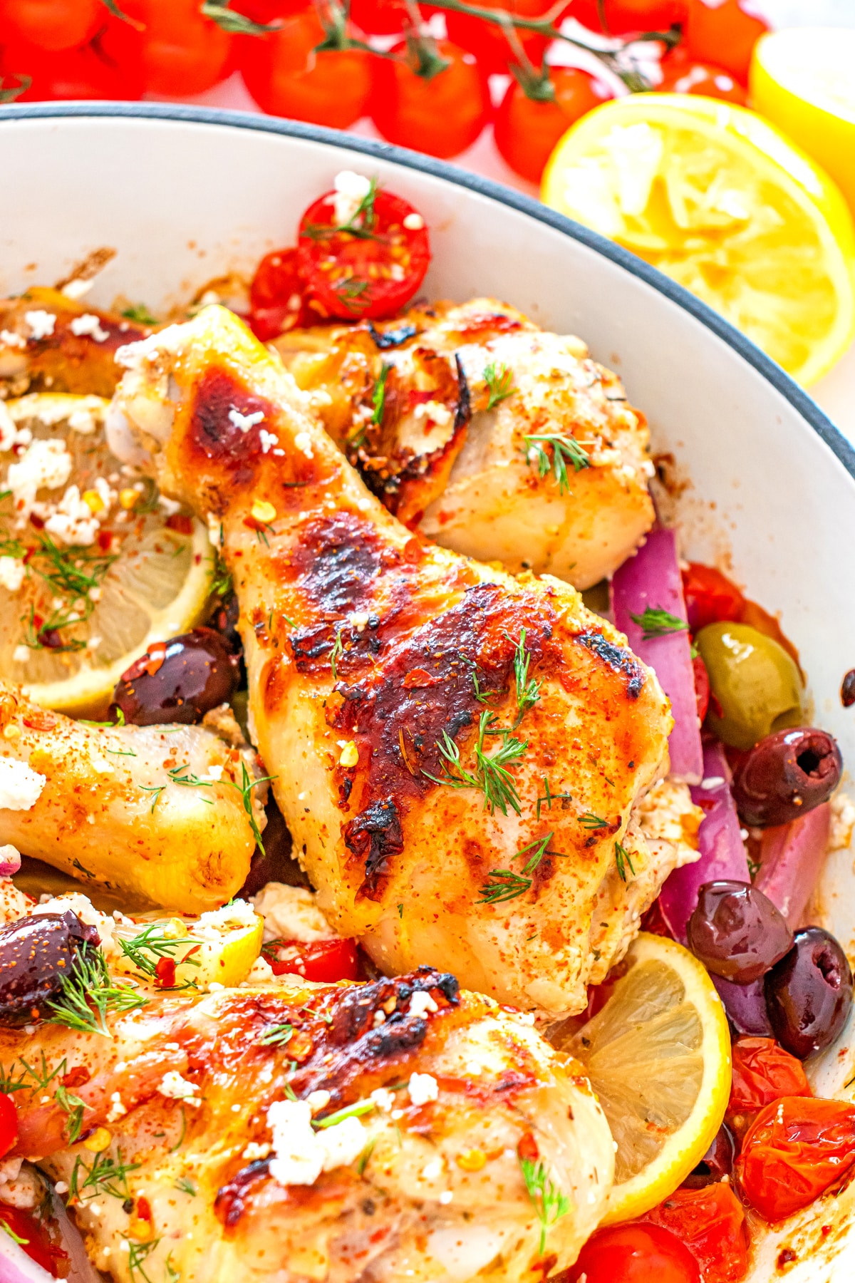 A closer look on One Pan Greek Chicken Drumsticks with lemon slices, cherry tomatoes, olives, red onions, herbs, and feta cheese in a white dish.