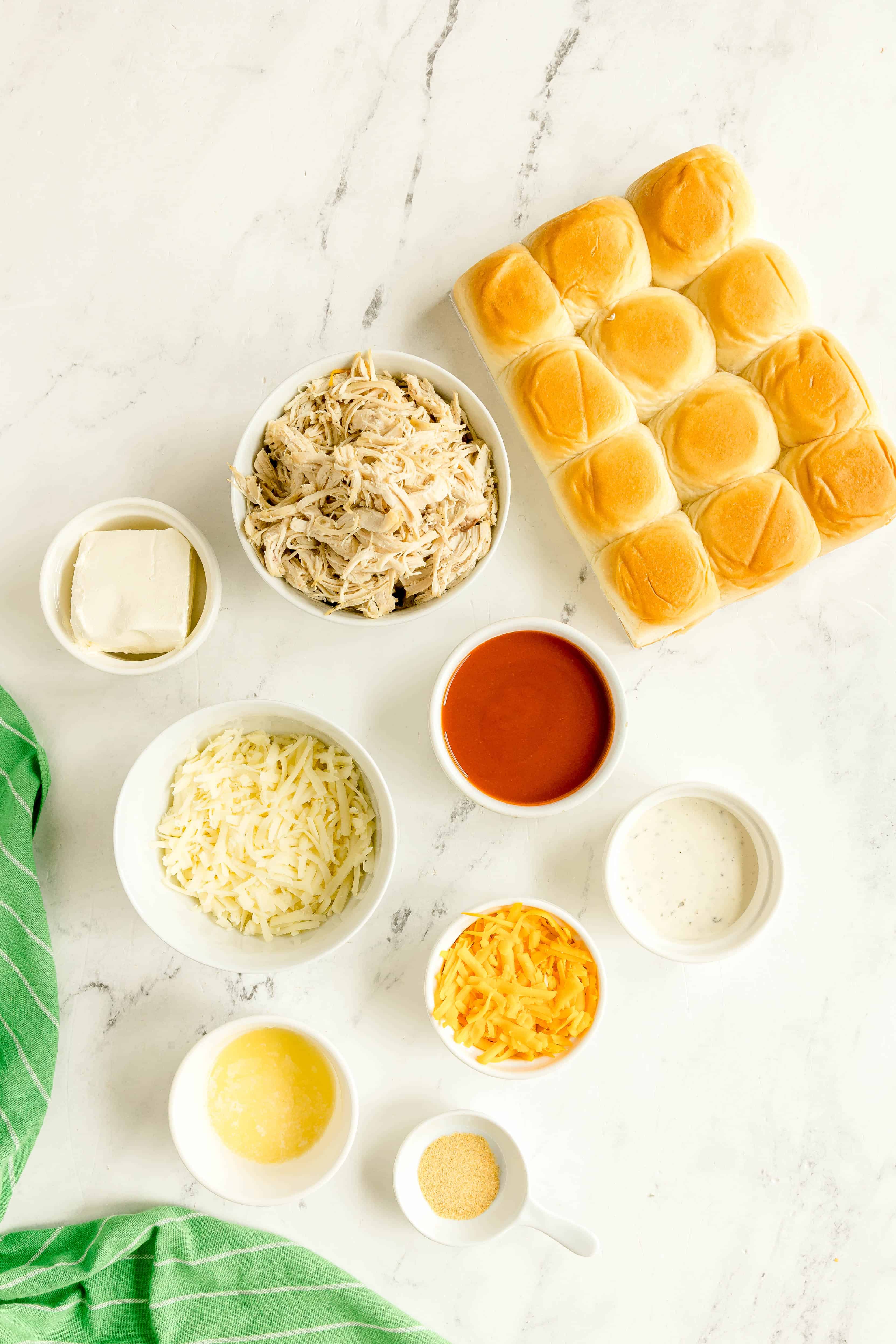 Top-down view of ingredients of Buffalo Chicken Sliders on a white surface including shredded chicken, Hawaiian sweet rolls, buffalo sauce, shredded cheese, cheddar cheese, cream cheese, ranch dressing, unsalted butter, and garlic powder on a white counter.