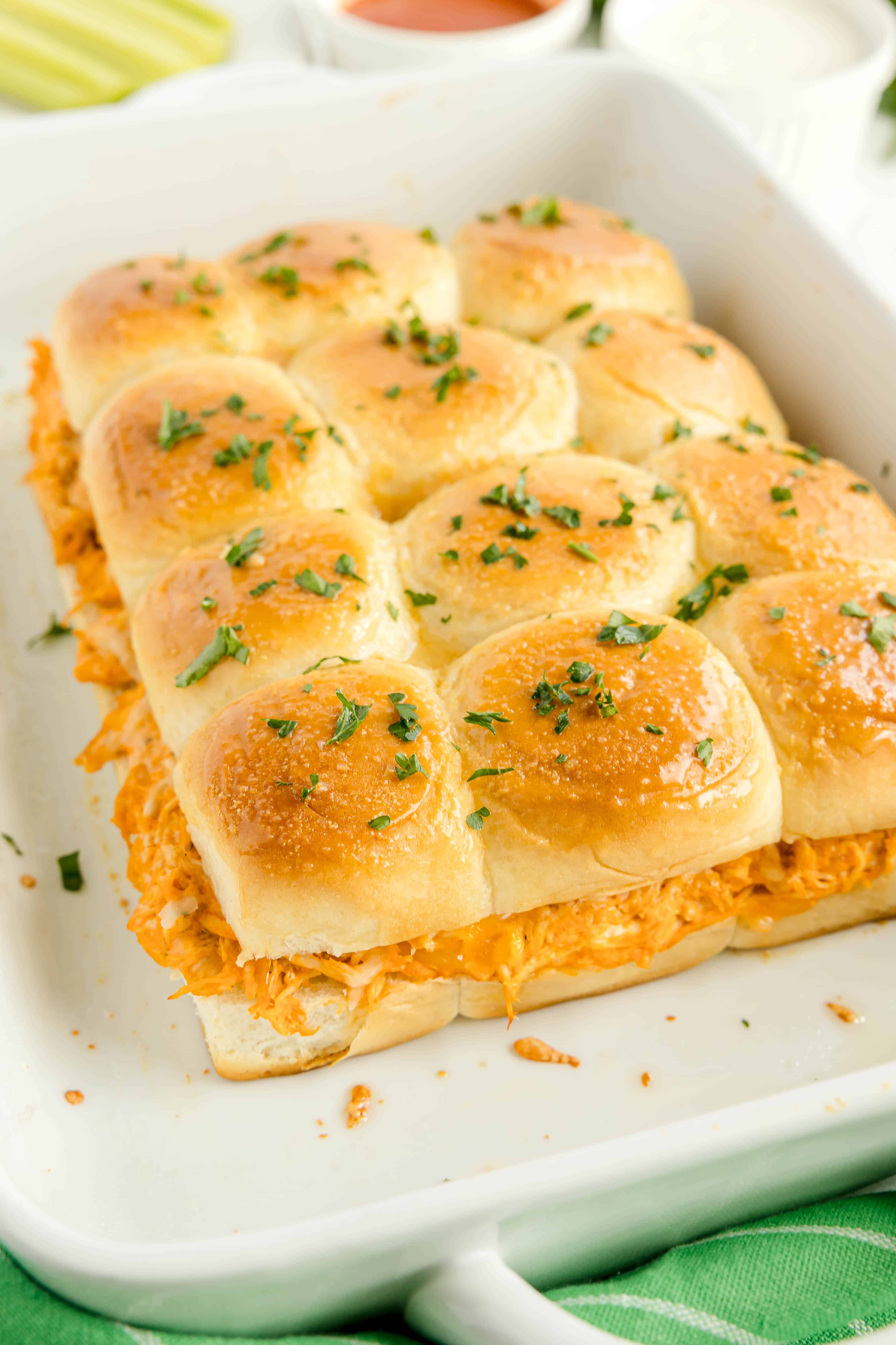 Golden slider rolls filled with shredded buffalo chicken, topped with chopped parsley, in a white baking dish.