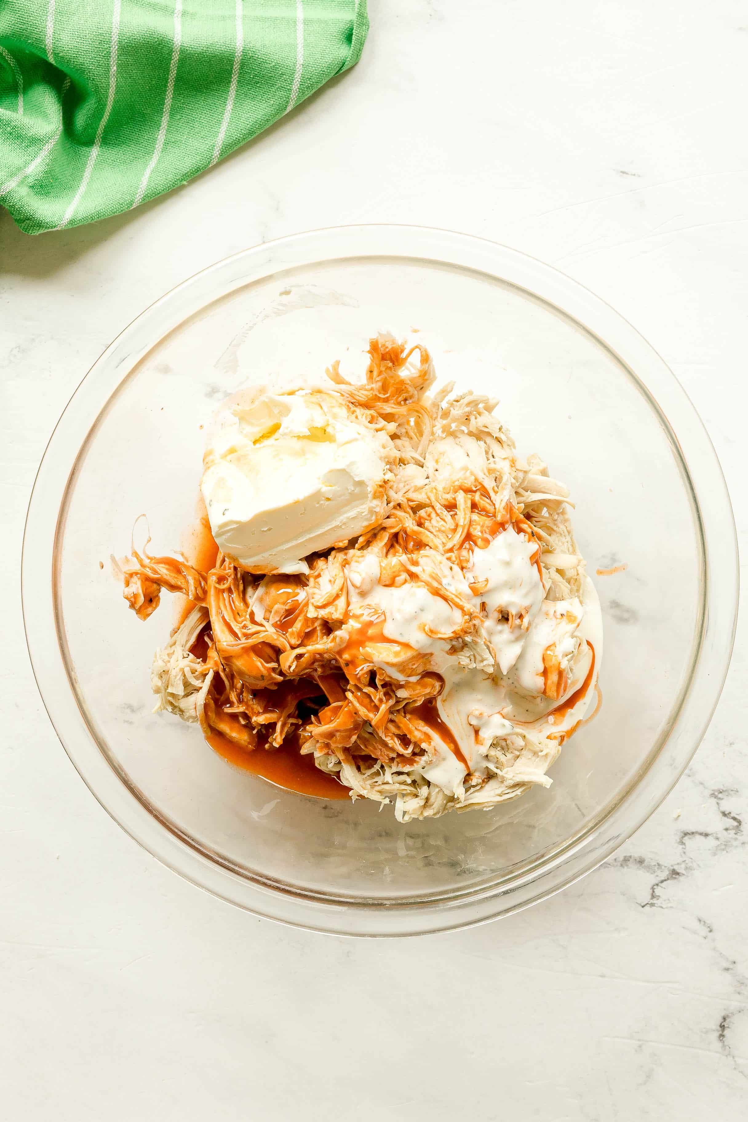 A glass bowl containing shredded chicken, buffalo sauce, softened cream cheese, and ranch dressing mixture, on a white surface with a green striped cloth nearby.