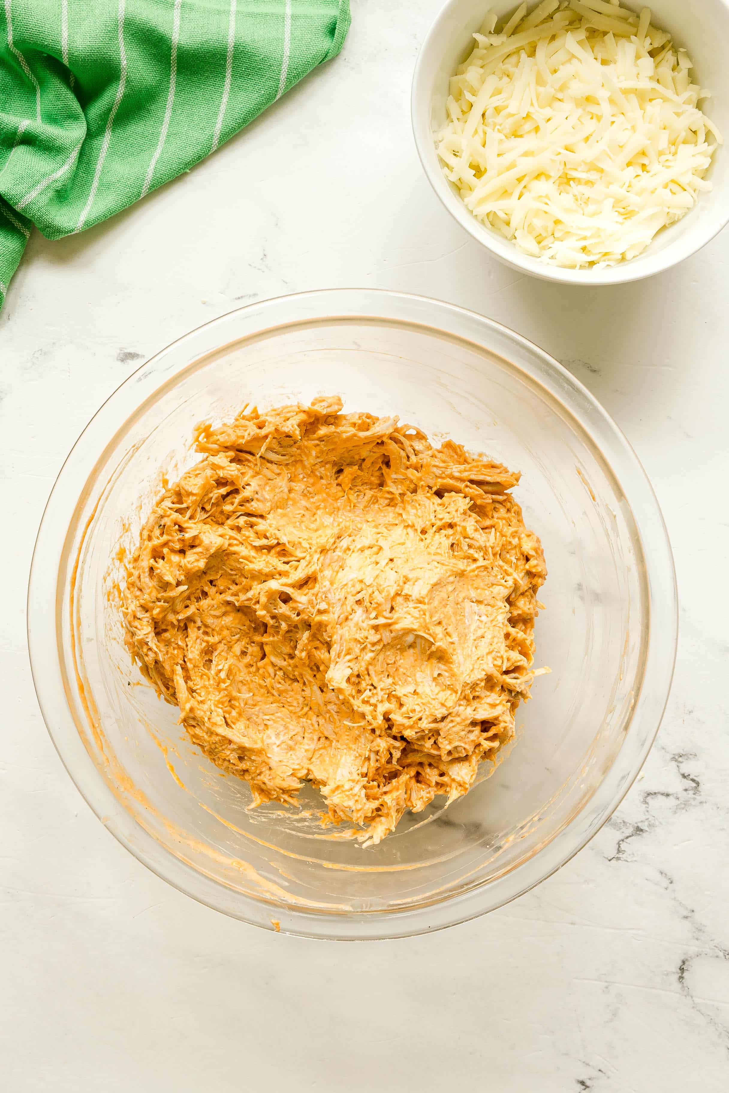 A glass bowl with a creamy orange mixture of mozzarella and part of the cheddar cheese, sits on a white countertop next to a bowl of shredded cheese and a green striped cloth. This is one of the steps in preparing Buffalo Chicken Sliders