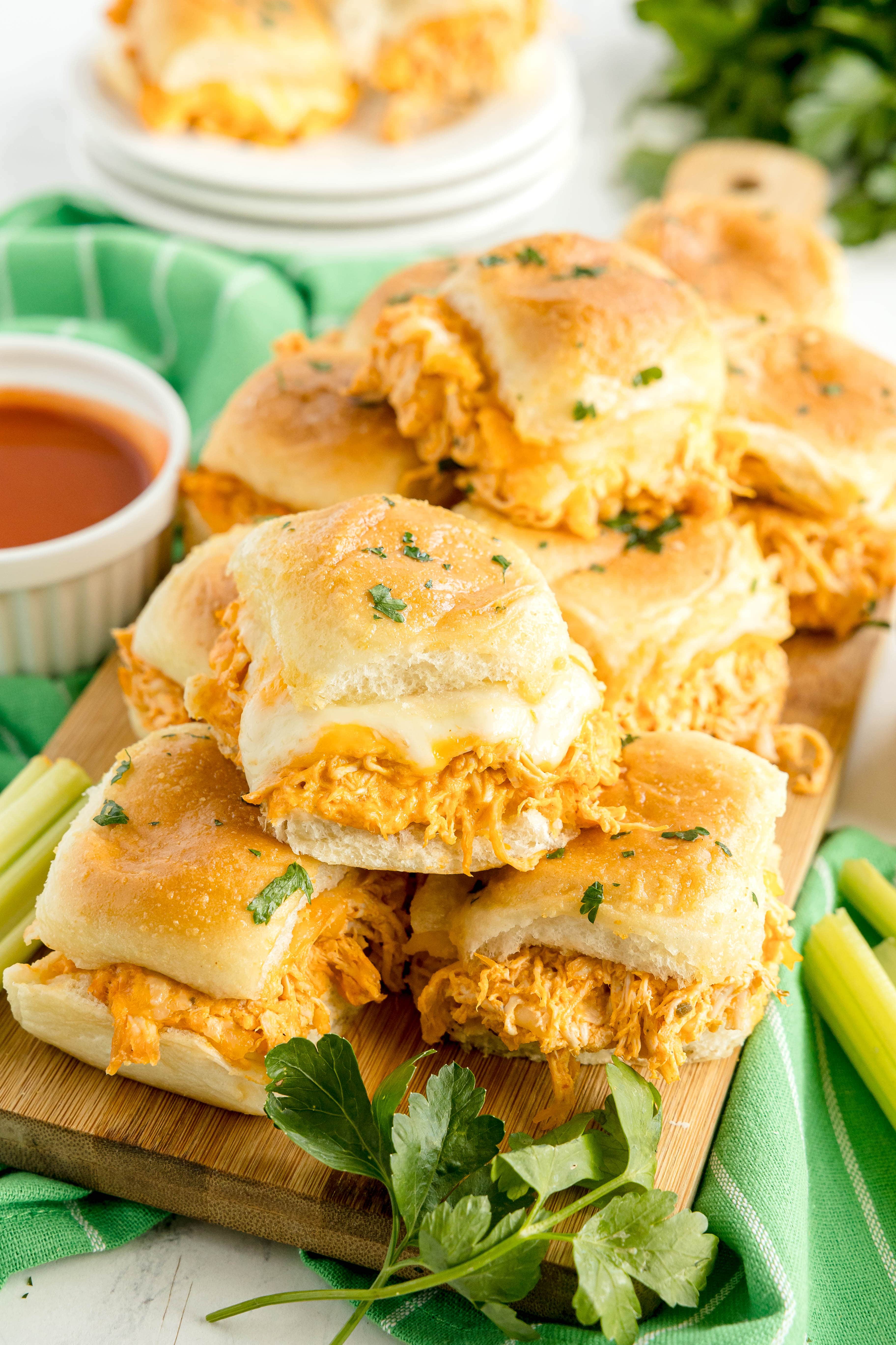 A wooden board topped with several buffalo chicken sliders, garnished with parsley, next to a ramekin of dipping sauce and celery sticks.