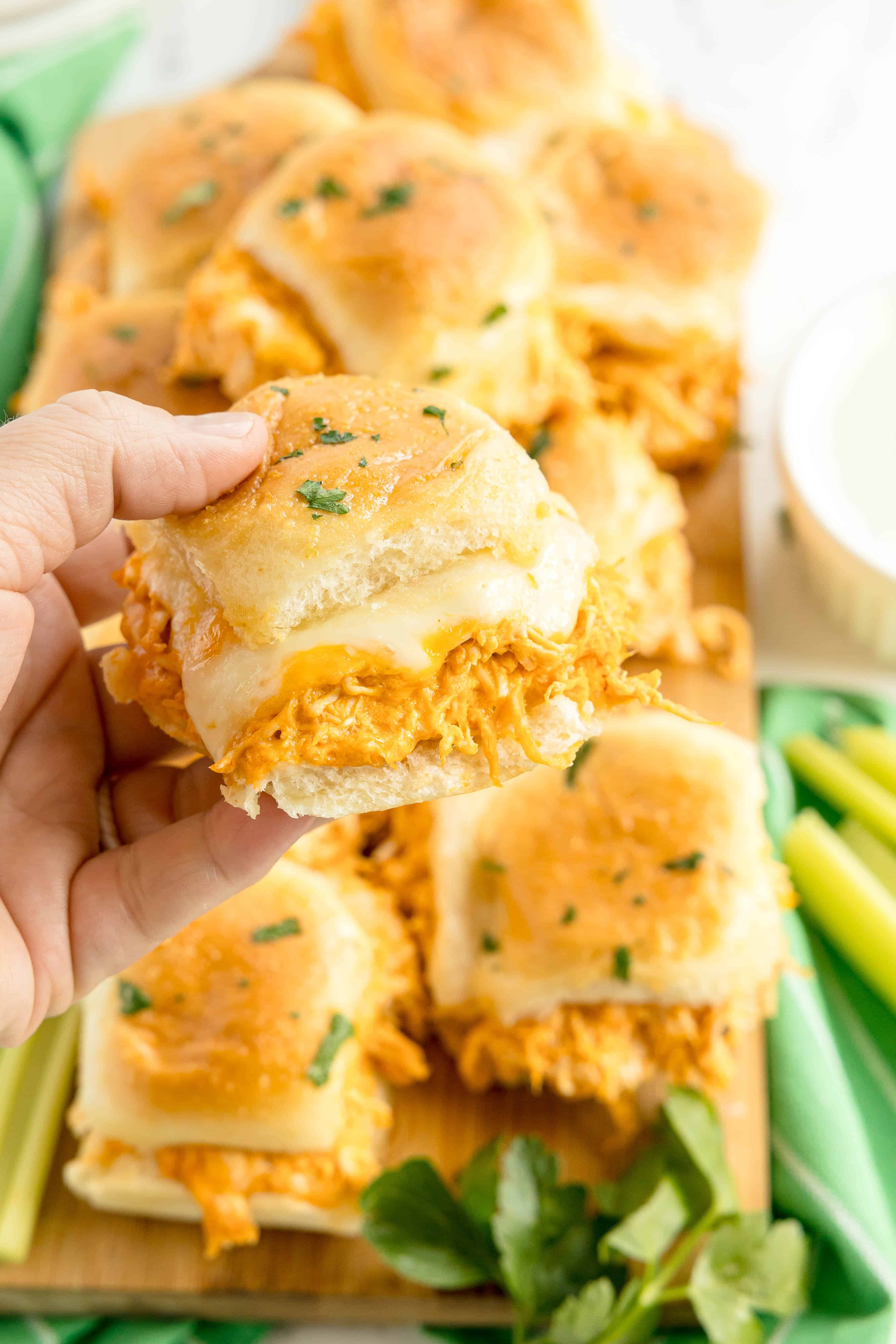A hand holds a slider sandwich filled with shredded chicken and melted cheese, with more sliders and fresh herbs visible on a wooden board in the background.