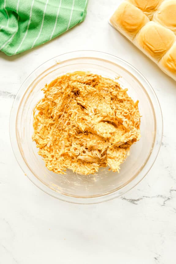 A glass bowl filled with a mixture of shredded chicken and sauce sits on a white surface, next to a green striped towel and a pack of bread rolls for Buffalo Chicken Sliders recipe.