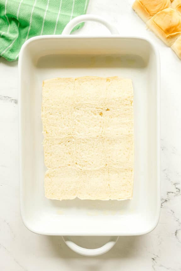 Next step in preparing Buffalo Chicken Sliders is to arrange a layer of plain, sliced bread rolls arranged in the bottom of a white rectangular baking dish on a marble surface.