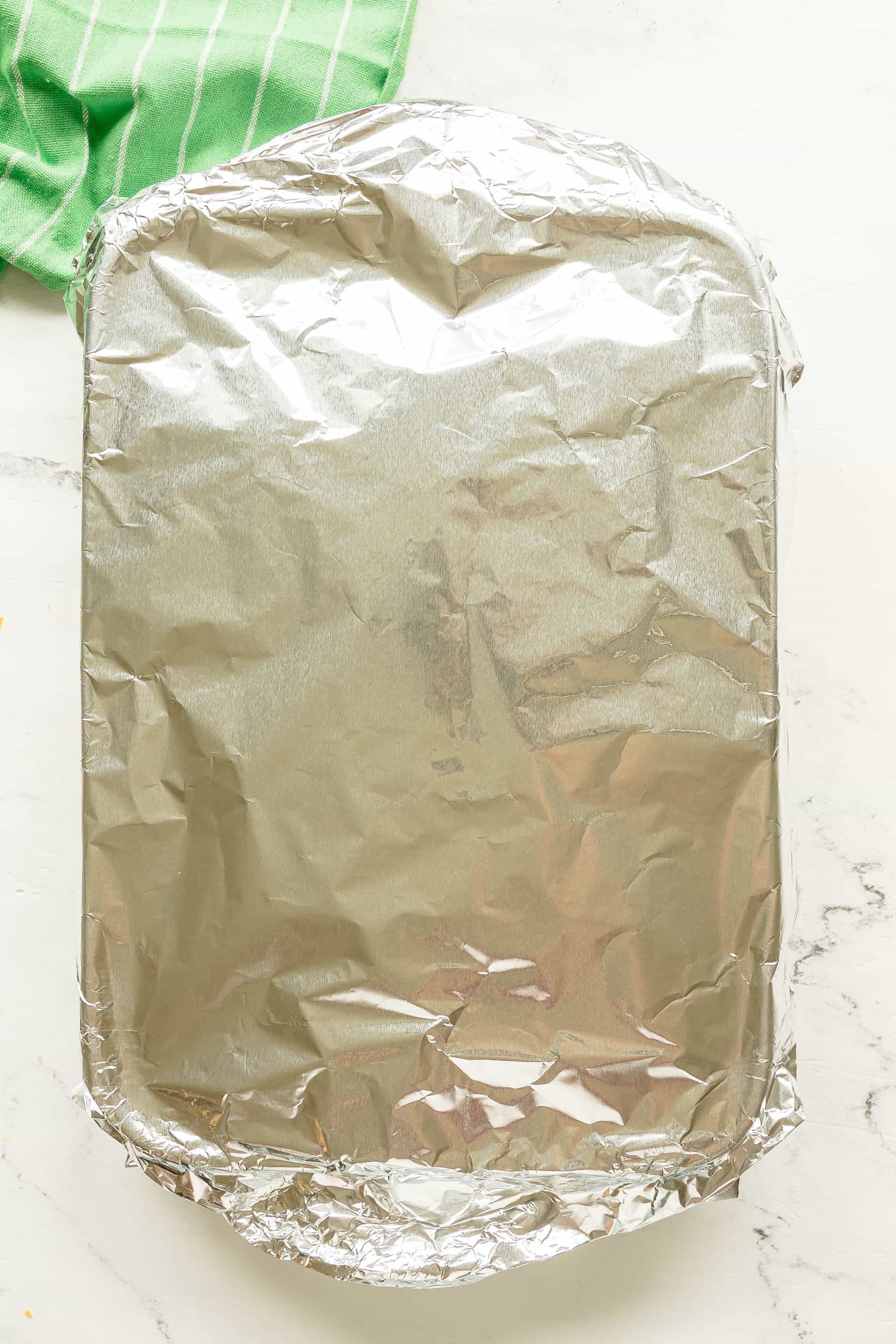 A rectangular baking dish of Buffalo Chicken Sliders, covered with aluminum foil sits on a white countertop next to a green striped cloth.