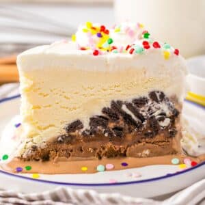 A slice of layered ice cream cake with chocolate and vanilla ice cream, cookie and graham crust, topped with whipped cream and colorful sprinkles on a white plate.