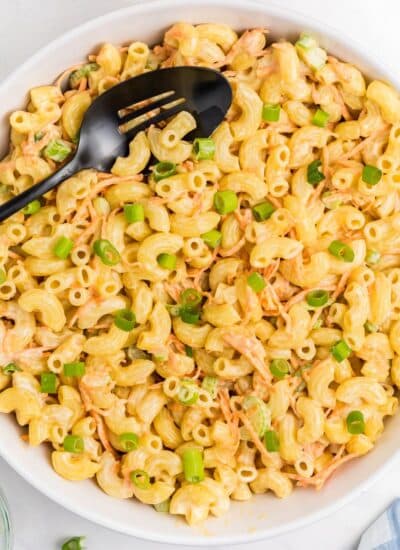 Bowl of macaroni Hawaiian salad with creamy dressing, shredded carrots, and chopped green onions, served with a black serving spoon.