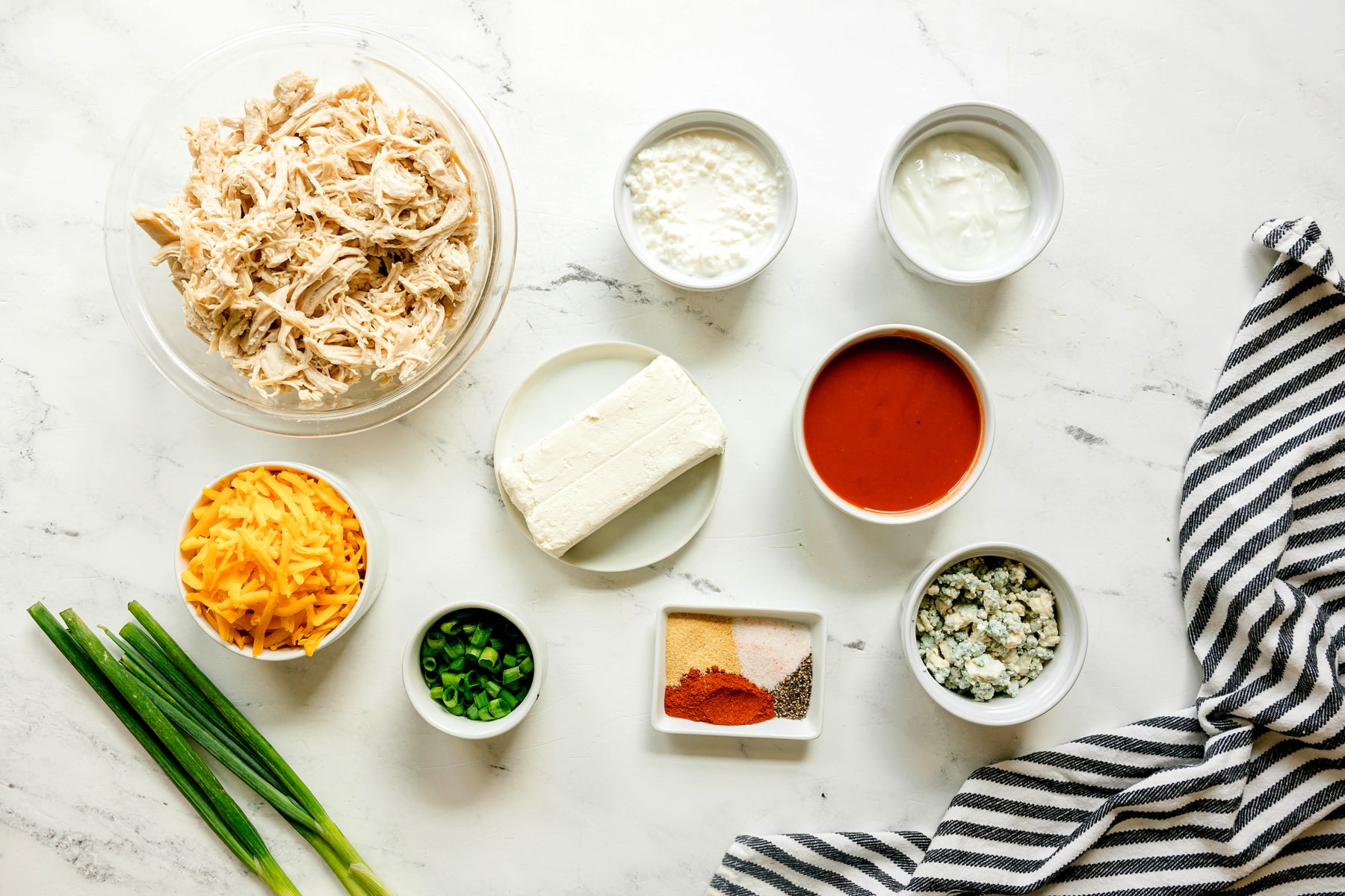 Ingredients for High Protein Buffalo Chicken Dip arranged on a white surface: shredded Rotisserie chicken, cream cheese, cheddar, green onions, cottage cheese, Greek yogurt, Frank's RedHot sauce, garlic powder, smoked paprika, salt, black pepper, blue cheese, and scallions.