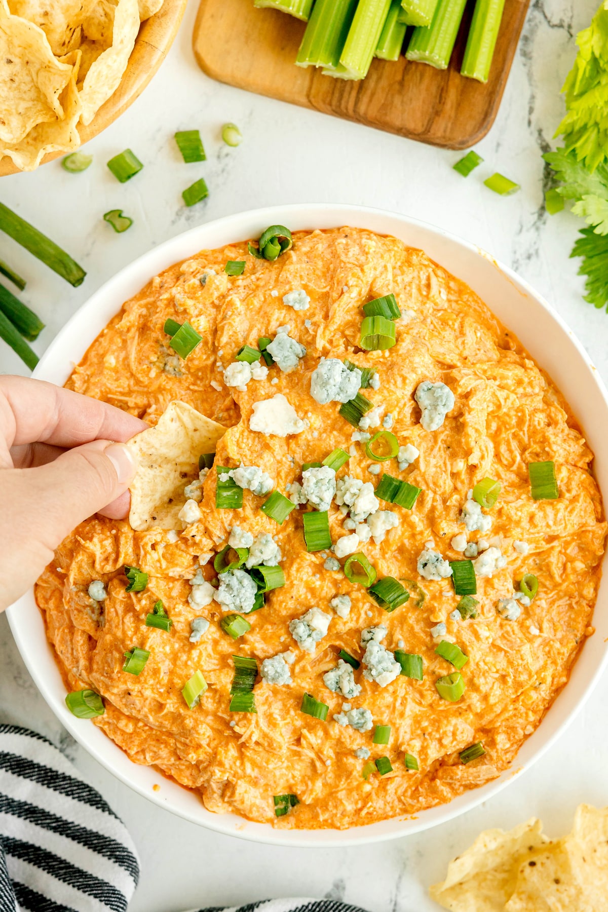 A hand dips a tortilla chip into a bowl of High Protein Buffalo Chicken Dip, topped with blue cheese crumbles and chopped green onions. Celery sticks and chips are visible nearby.