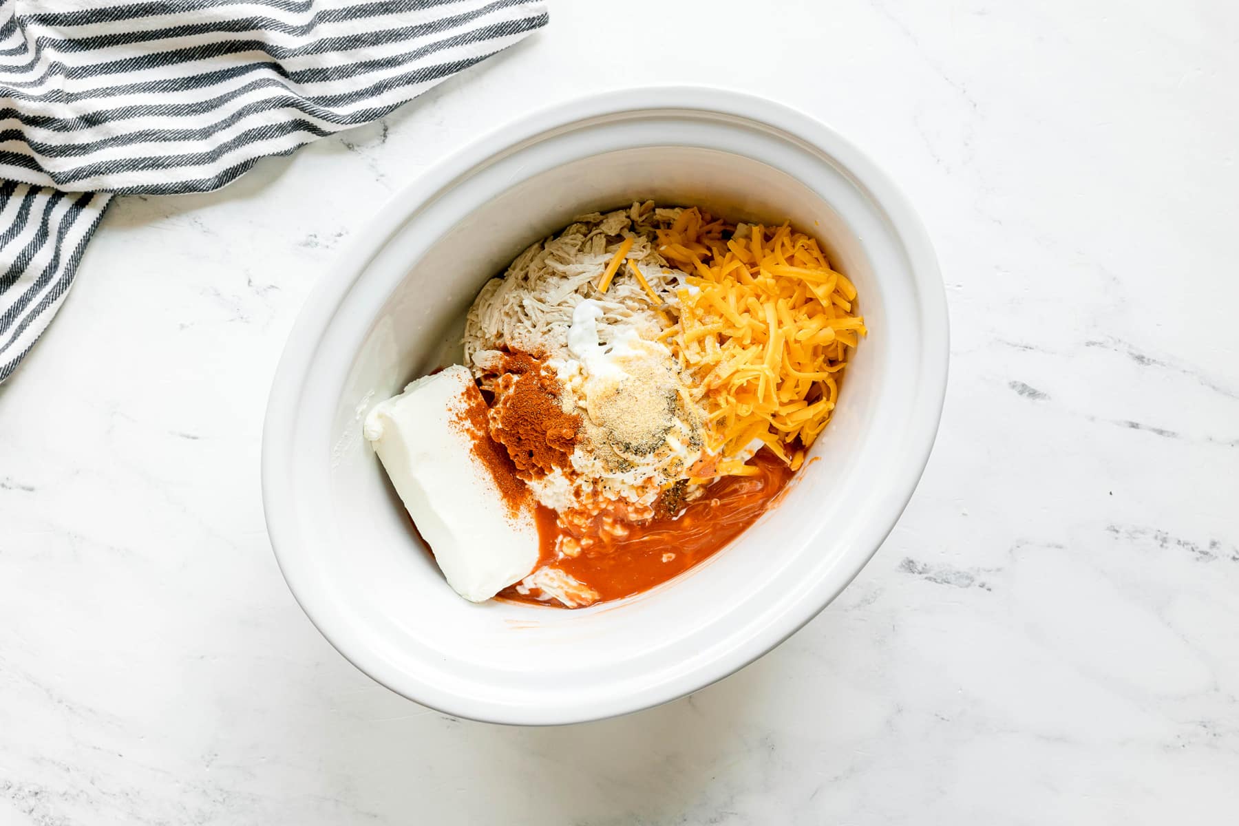 A white slow cooker filled with ingredients for High Protein Buffalo Chicken Dip. It includes: chicken, cottage cheese, Greek yogurt, cream cheese, cheddar, Frank's hot sauce, garlic powder, smoked paprika, salt, and pepper on a marble countertop.